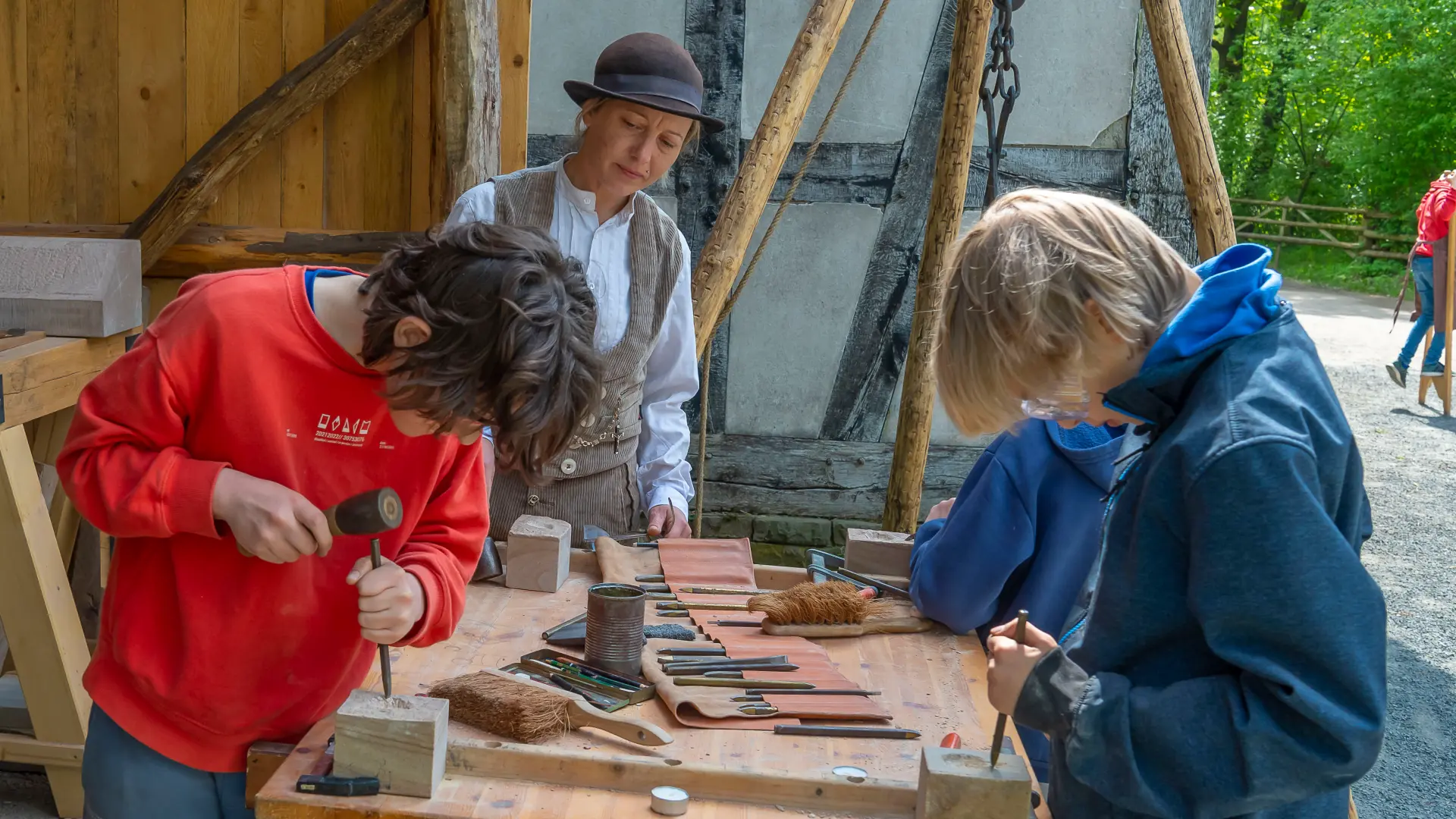 Foto: Drei Personen an einer Werkbank vor einem historischen Fachwerkgebäude. Zwei Kinder bearbeiten mit Hammer und Werkzeugen kleine Werkstücke aus Stein oder Holz. Eine erwachsene Person steht dahinter und begleitet die Arbeit.