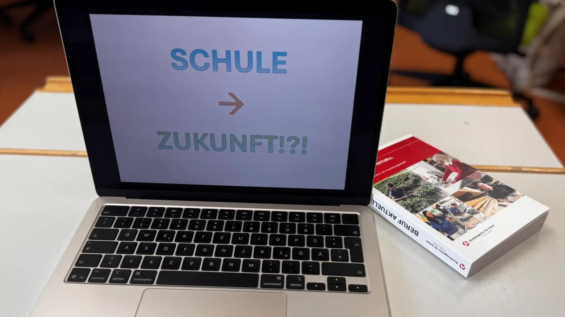 Ein aufgeklappter Laptop mit dem Schriftzug 'Schule > Zukunft?' am Monitor, dazu ein Buch mit dem Titel 'Aktuelle Berufswelten'
