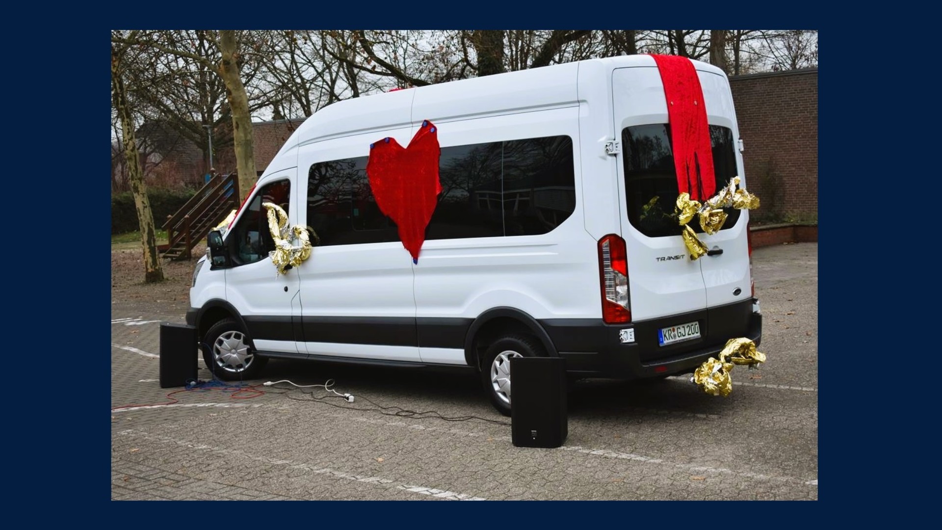 Ein weißer Kleintransporter ist mit Goldfolie und roten Herzen geschmückt. An zwei seiner Räder steht jeweils eine Lautsprecherbox.