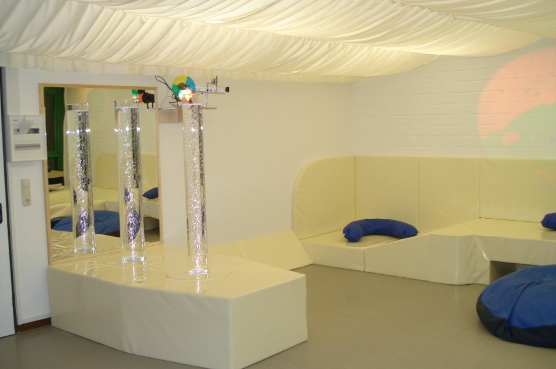 Ein Snoezelen-Raum zur Entspannung: Weiße Polsterecken und blaue Sitzsäcke laden zum Verweilen ein. Sprudelnde, beleuchtete Wassersäulen vor Spiegeln und Lichtprojektionen bieten sanfte Sinnesreize.