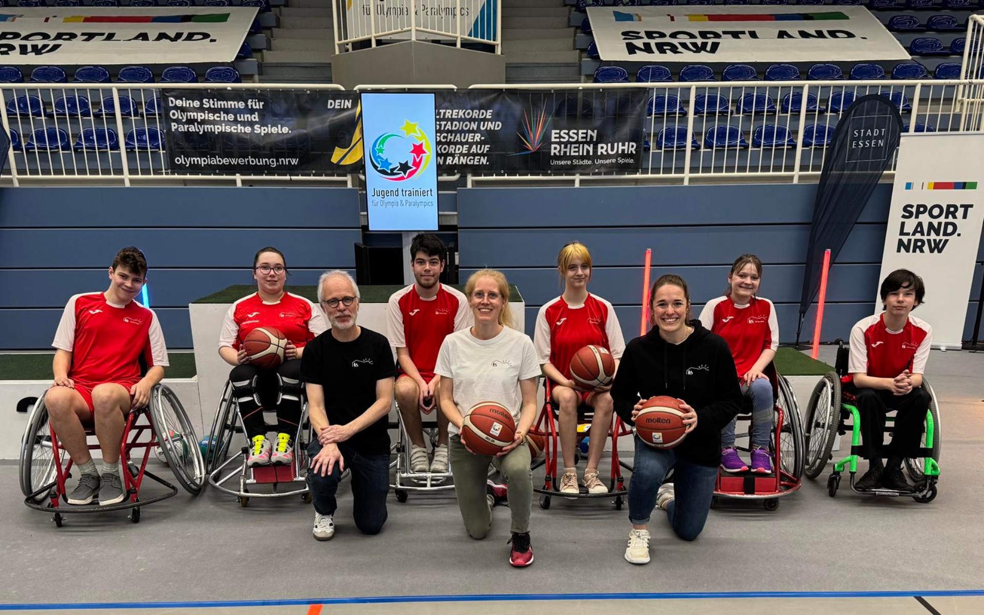 Ein Team aus Rollstuhl-Basketball-Spieler*innen mit roten Krefels-Trikots und deren TrainerTrainerinnen im Kreis.