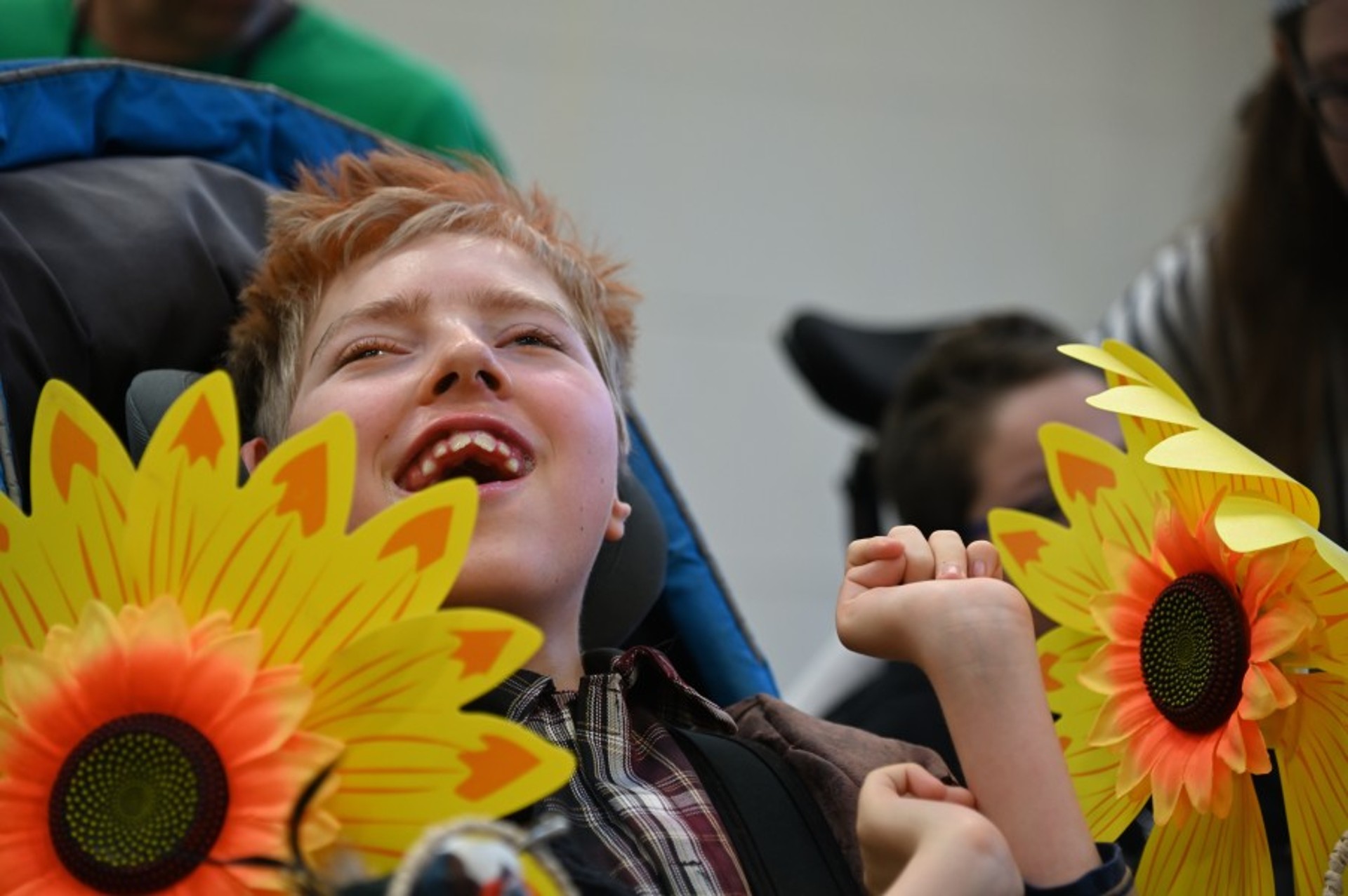 Gesicht eines frohen Kindes im Rollstuhl, an dem rechts und links große Blüten von Sonnenblumen befestigt sind.