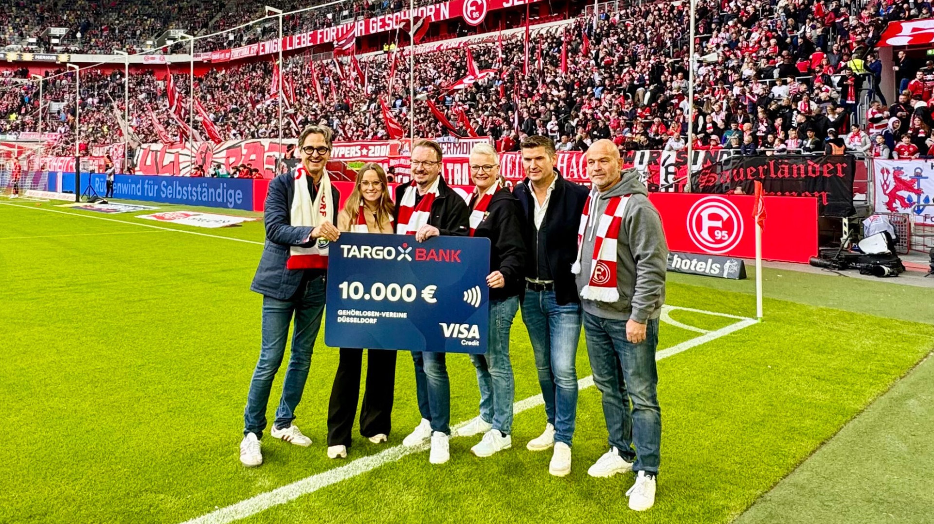 Eine Spendenübergabe im Fußballstadion von Fortuna Düsseldorf: Eine Gruppe von sechs Personen steht auf dem Rasen und hält einen symbolischen Scheck der Targobank über 10.000 € in die Kamera. Der Scheck ist an die „Gehörlosen-Vereine Düsseldorf“ adressiert. Im Hintergrund sind die vollbesetzten Tribünen mit Fans in rot-weißer Fankleidung zu sehen.