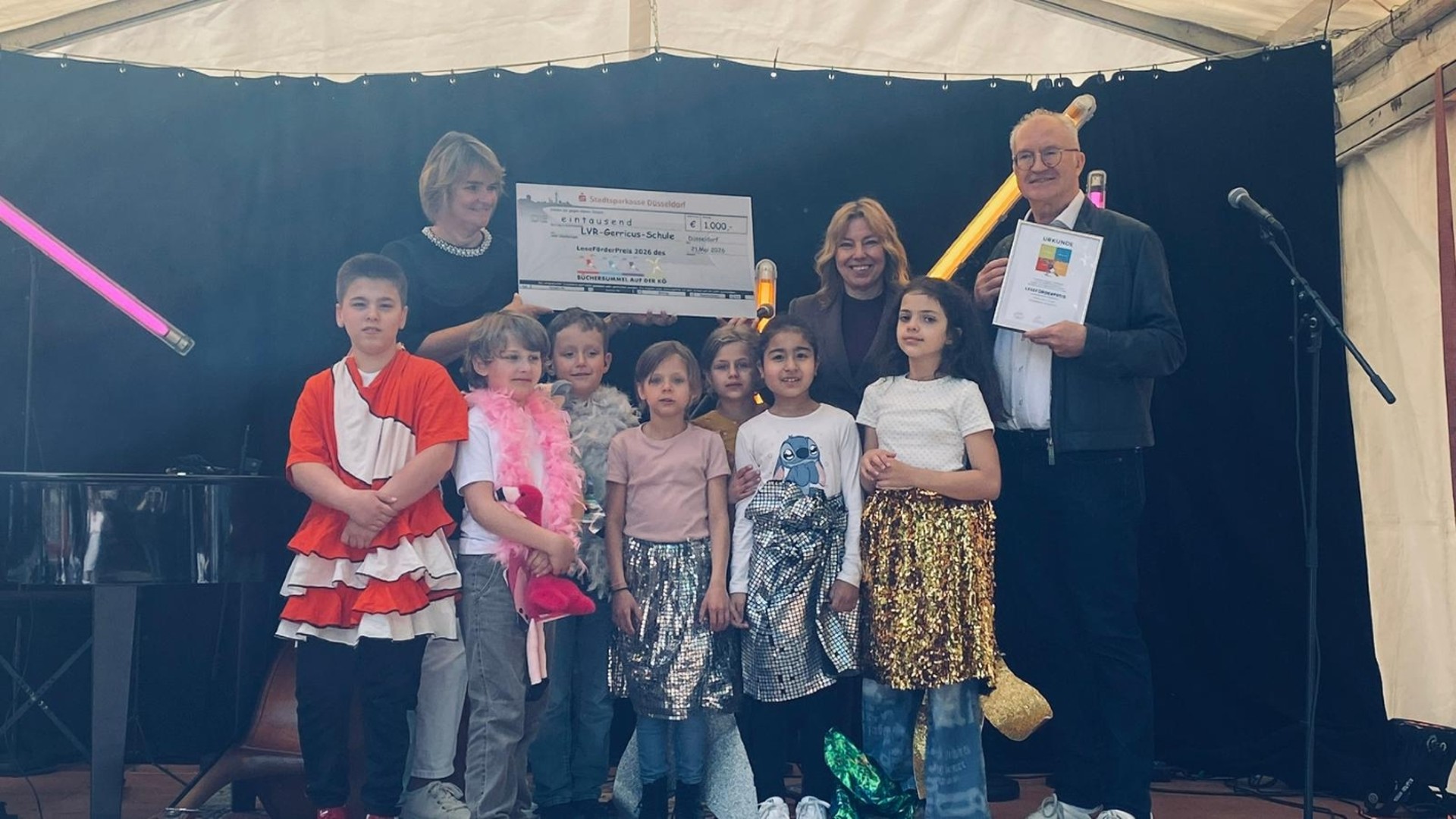 Eine Gruppe von sieben Personen steht vor der Schulmauer und hält einen symbolischen Scheck der Stadtsparkasse Düsseldorf über 1000 € in die Kamera. Der Scheck ist an die „LVR-Gerricus-Schule“ adressiert. Im Hintergrund sind Bäume, die bemalte Schulmauer und ein Teil des Schulgebäudes zu sehen.