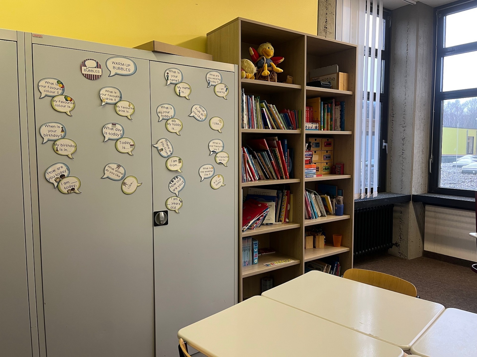 In einem Raum steht an einer gelben Wand ein grauer Metallschrank. Auf ihm kleben Sprechblasen mit englischen Fragen und dem Beginn möglicher Antworten (Warm-up-bubbles), zum Beispiel "How are you today?" – "I am ... today." Daneben steht ein Regal mit Büchern und weiteren Unterrichtsmaterialien.