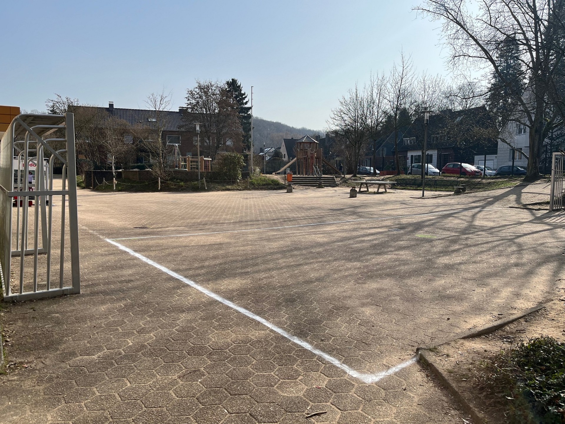 Großer, gepflasterter Pausenhof der Grundschule mit aufgemalten Spielfeldern, einem Klettergerüst im Hintergrund und angrenzenden Wohnhäusern.