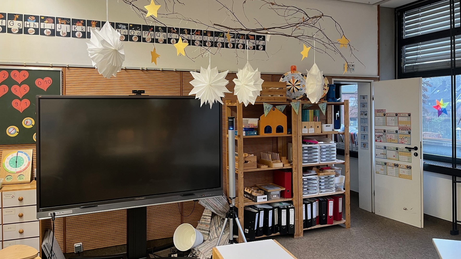 Ein gemütlicher Lernbereich mit einem digitalen Whiteboard und Regalsystemen. Ein großer Naturast, der mit Papiersternen und -lampen dekoriert ist, hängt von der Decke.