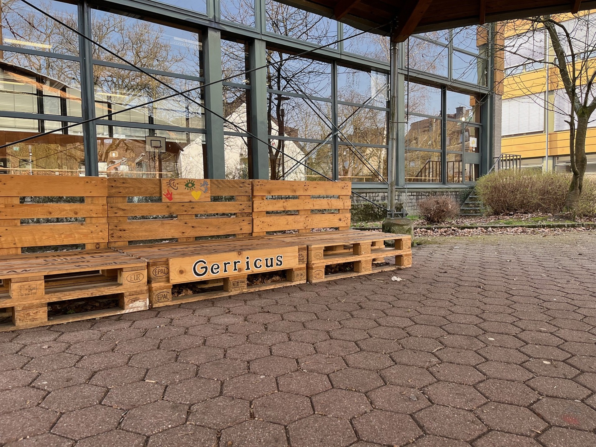 Detailaufnahme einer aus Europaletten gefertigten Sitzbank mit der Aufschrift „Gerricus“ und bunten Symbolen vor der Glasfassade der Schulsporthalle.