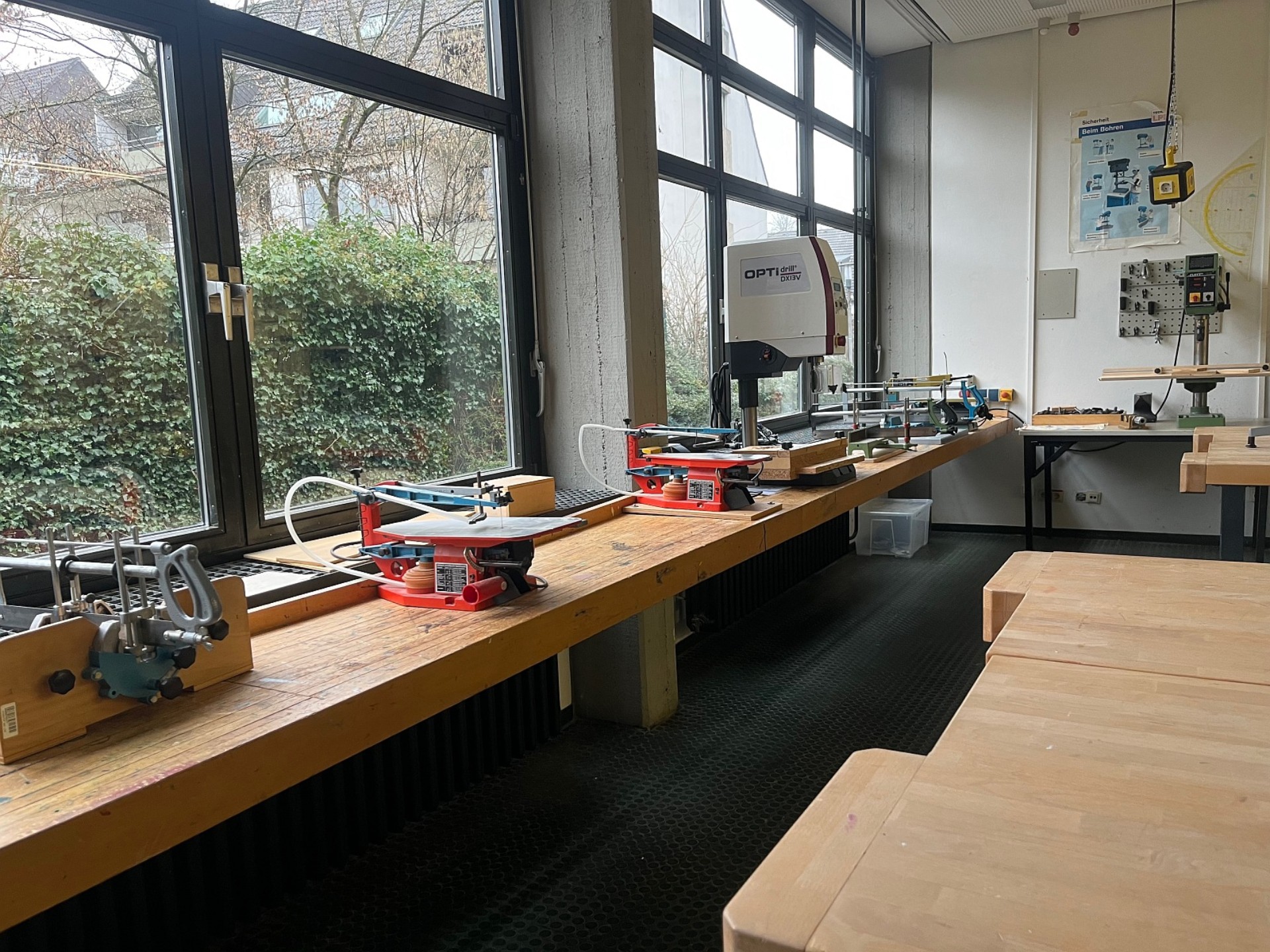 Eine Werkstatt mit einer langen Massivholz-Werkbank vor einer Fensterfront, auf der mehrere Dekupiersägen und eine Standbohrmaschine montiert sind. Im Vordergrund sind schwere Hobelbänke sichtbar.