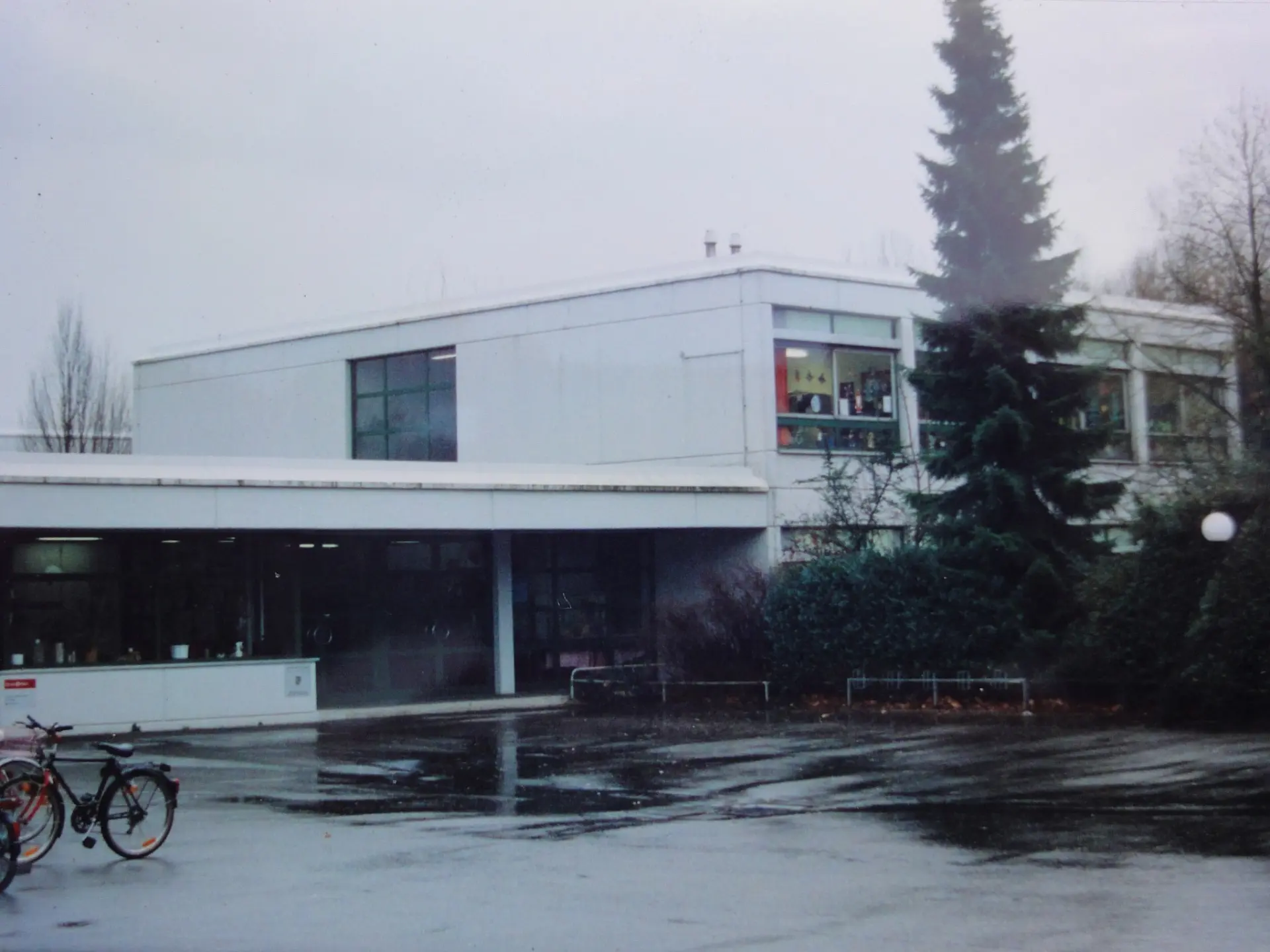 Ansicht des ehemaligen Schulgebäudes der Heinrich-Welsch-Schule im Stadtteil Flittard, ein funktionaler Bau mit flachem Dach und einem großen Tannenbaum im Vordergrund.