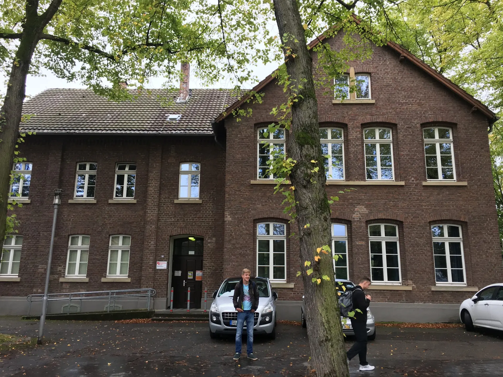 Außenansicht des historischen Backsteingebäudes der ehemaligen Hauptschule in Köln-Holweide mit großen Fenstern und einem hohen Giebel, eingerahmt von grünen Bäumen.