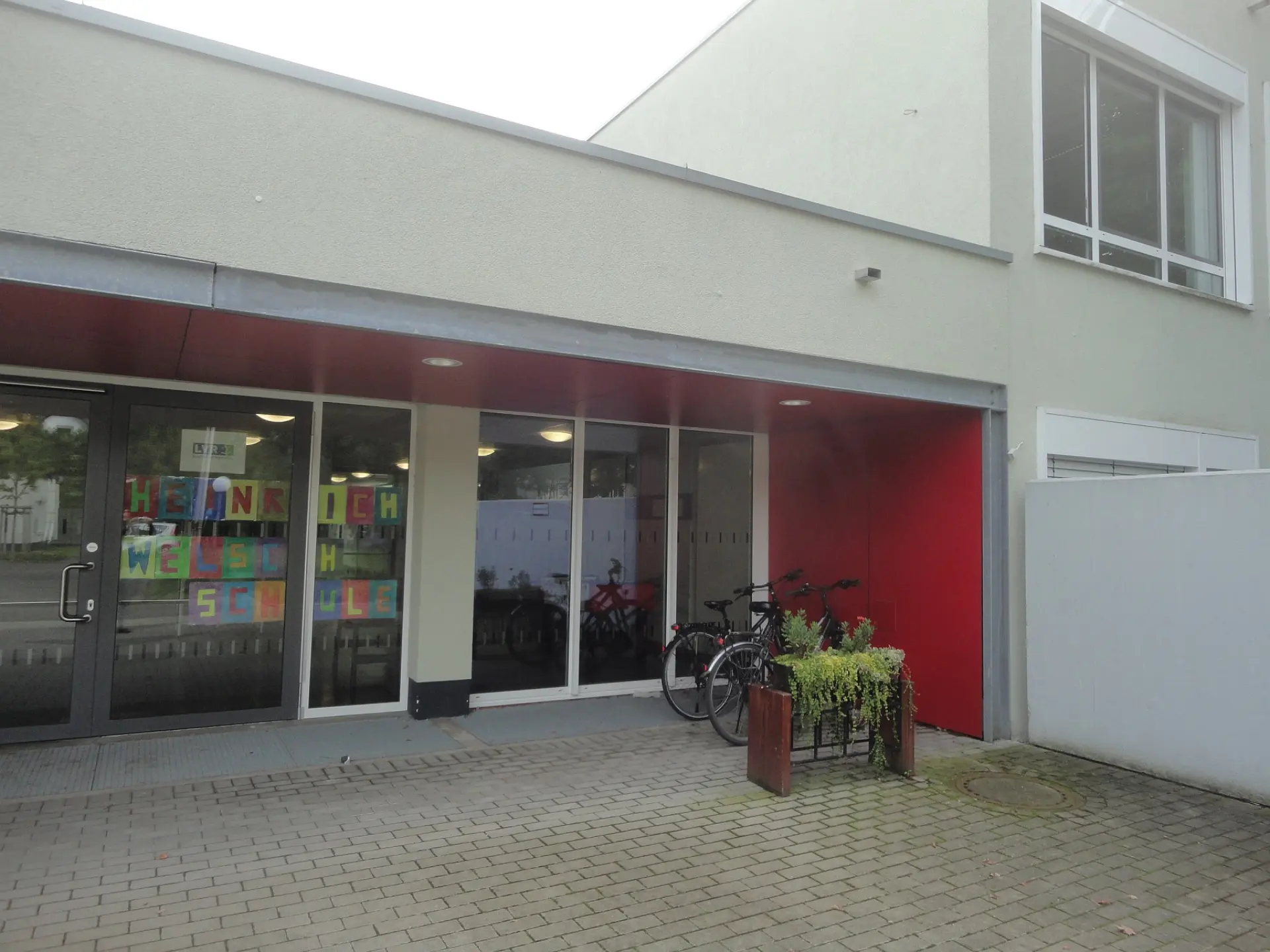 Der modernisierte Eingangsbereich der LVR-Heinrich-Welsch-Schule mit einer hellen Fassade, roten Akzenten an der Decke des Vorbaus und bunt gestalteten Buchstaben im Fenster, die den Schulnamen bilden.
