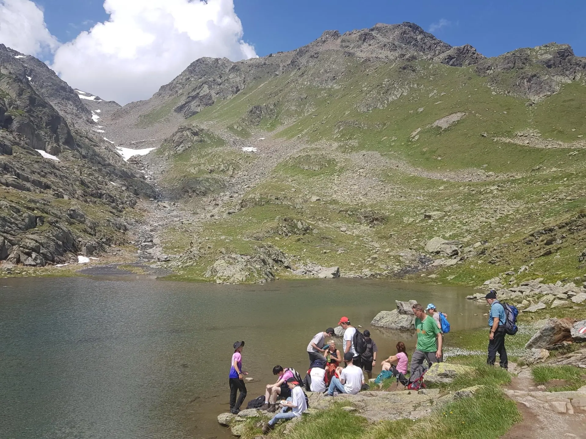Eine große Gruppe aus Schüler*innen und ein paar Lehrkräften macht eine Wanderung in der Natur und läuft auf einem Weg an einem Fluss entlang. Alles drum-herum ist grün, das Wetter ist sonnig.