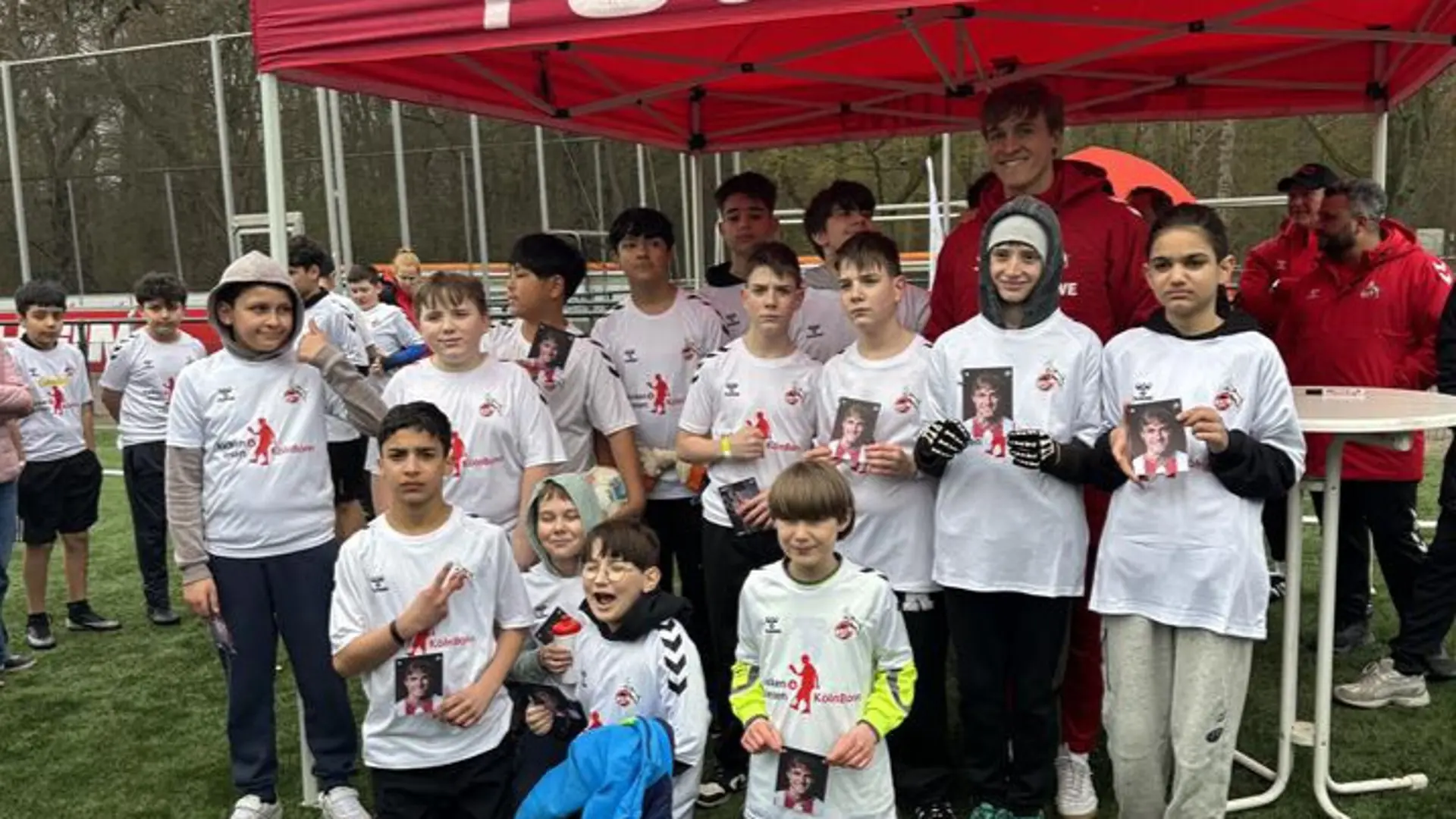 Ein Pavillon der FC Stiftung am Rande des Fußballspielplatzes. Davor stehen mehrere Kinder, alle mit weißen FC-Köln-Tshirts angezogen, halten eine Autogrammkarte in der Hand und schauen in die Kamera. Zu ihnen steht auch der FC-Köln-Profifußballer Timo Hübers und lächelt auch in die Kamera.