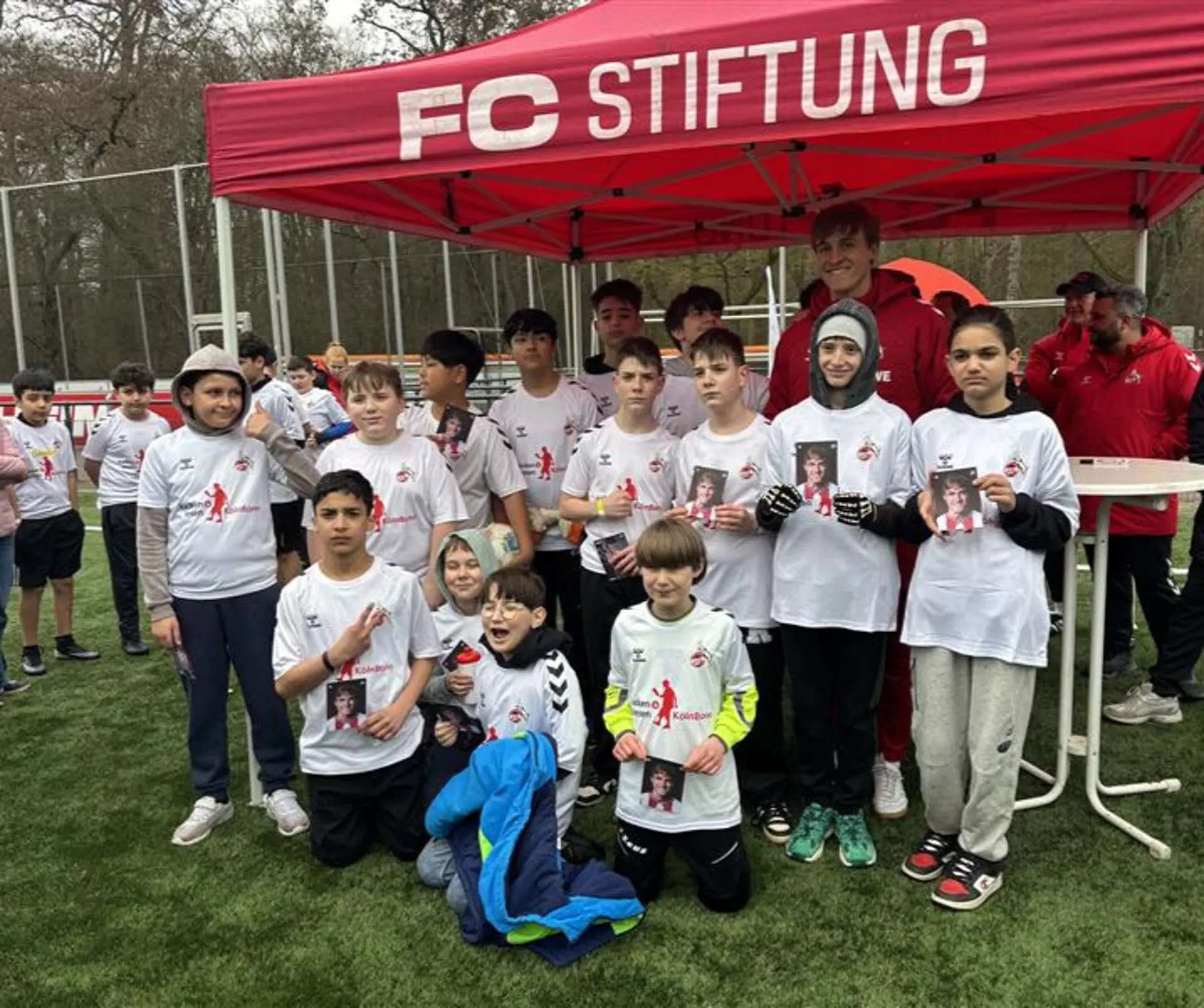 Ein Pavillon der FC Stiftung am Rande des Fußballspielplatzes. Davor stehen mehrere Kinder, alle mit weißen FC-Köln-Tshirts angezogen, halten eine Autogrammkarte in der Hand und schauen in die Kamera. Zu ihnen steht auch der FC-Köln-Profifußballer Timo Hübers und lächelt auch in die Kamera.
