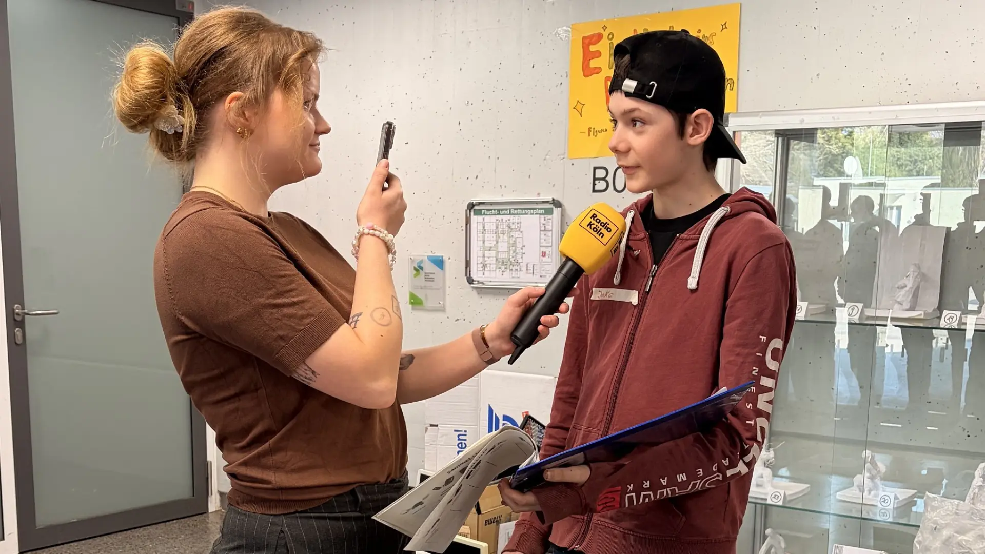 Eine Reporterin interviewt im Schulflur einen Schüler. In der einen Hand hält sie ein Mikrofon, das zum Schüler gerichtet ist, in der anderen ein Smartphone, mit dem sie ihre Stimme und Fragen aufnimmt.