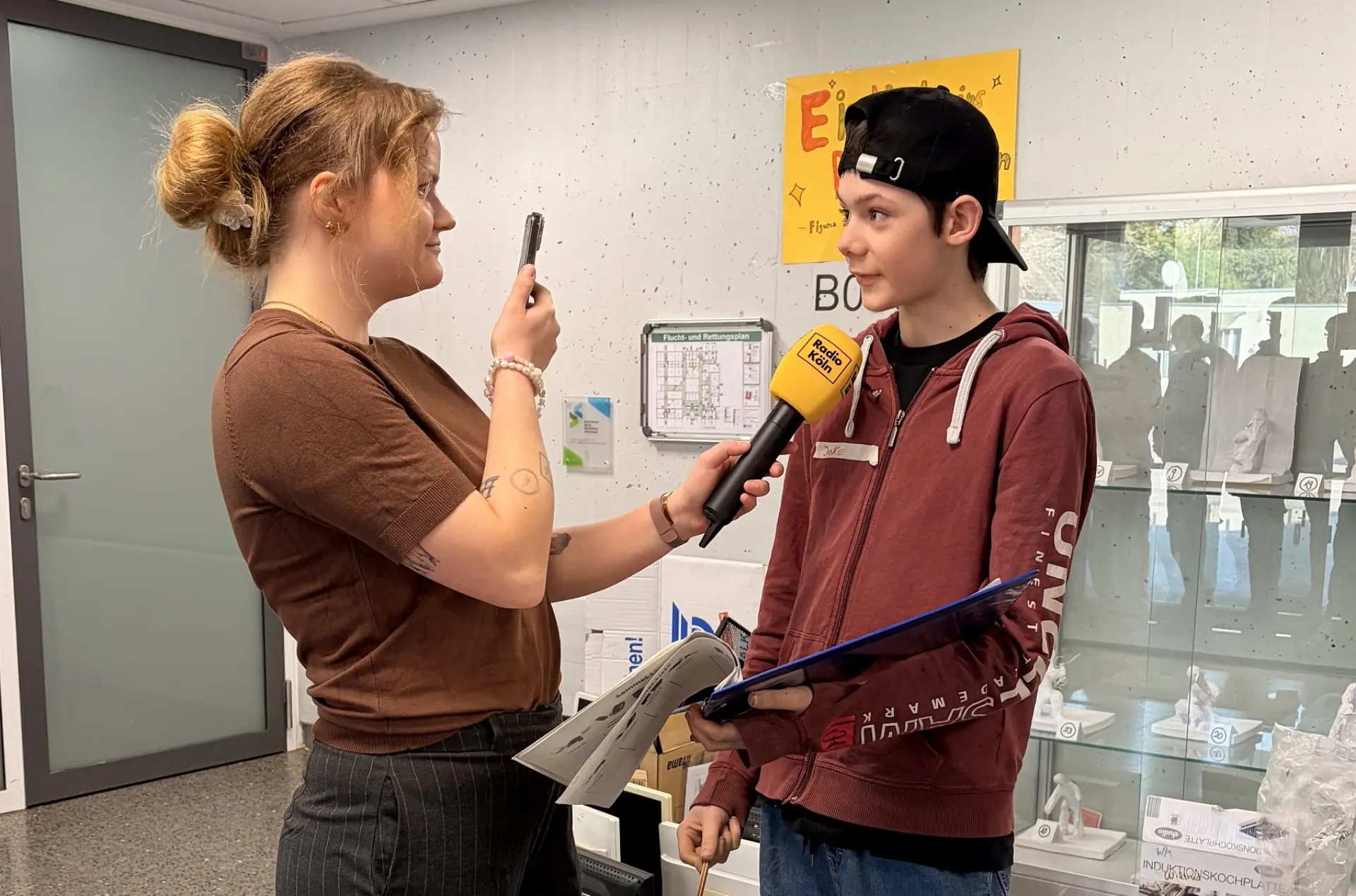 Eine Reporterin interviewt im Schulflur einen Schüler. In der einen Hand hält sie ein Mikrofon, das zum Schüler gerichtet ist, in der anderen ein Smartphone, mit dem sie ihre Stimme und Fragen aufnimmt.