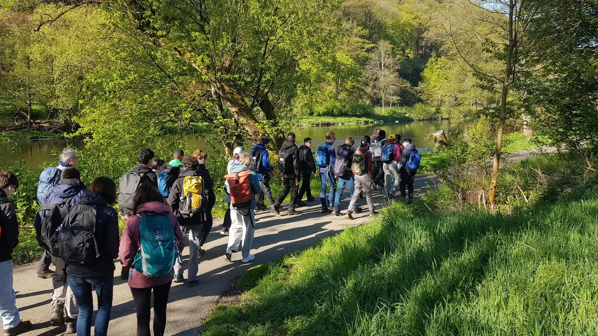 Eine große Gruppe aus Schüler*innen und ein paar Lehrkräften macht eine Wanderung in der Natur und läuft auf einem Weg an einem Fluss entlang. Alles drum-herum ist grün, das Wetter ist sonnig.