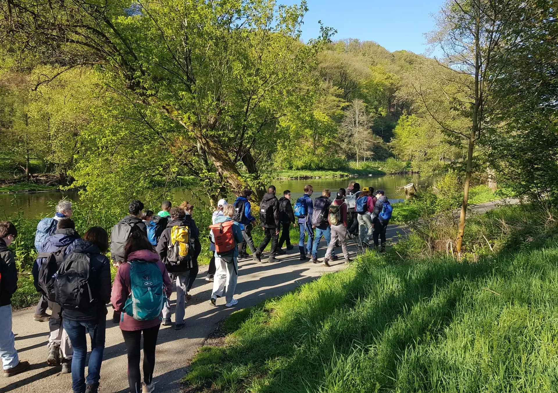 Eine große Gruppe aus Schüler*innen und ein paar Lehrkräften macht eine Wanderung in der Natur und läuft auf einem Weg an einem Fluss entlang. Alles drum-herum ist grün, das Wetter ist sonnig.