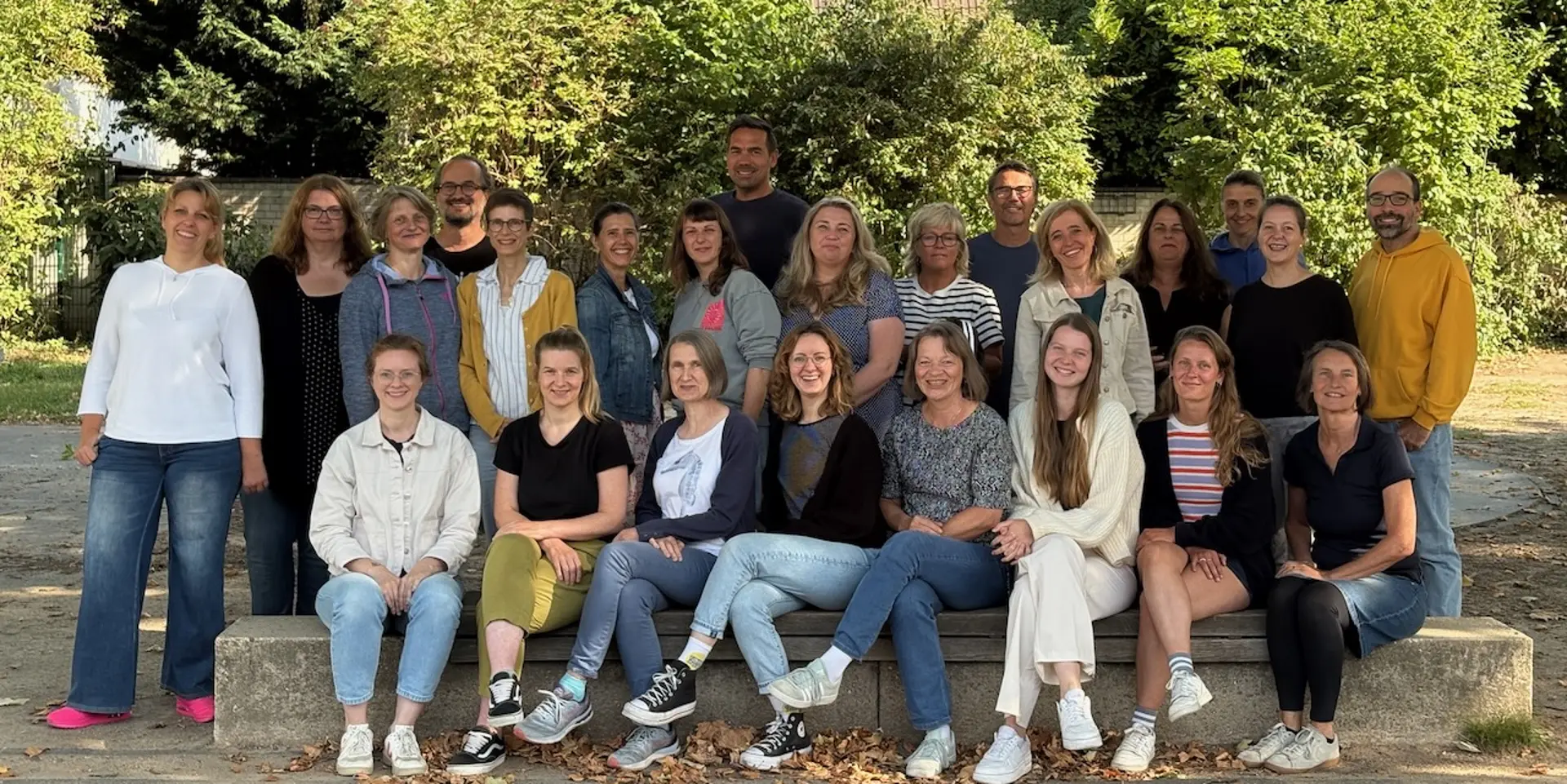 Eine Gruppe aus 24 Personen steht bzw. sitzt in zwei Reihen auf dem Schulhof. Sie alle schauen in die Kamera und lächeln.