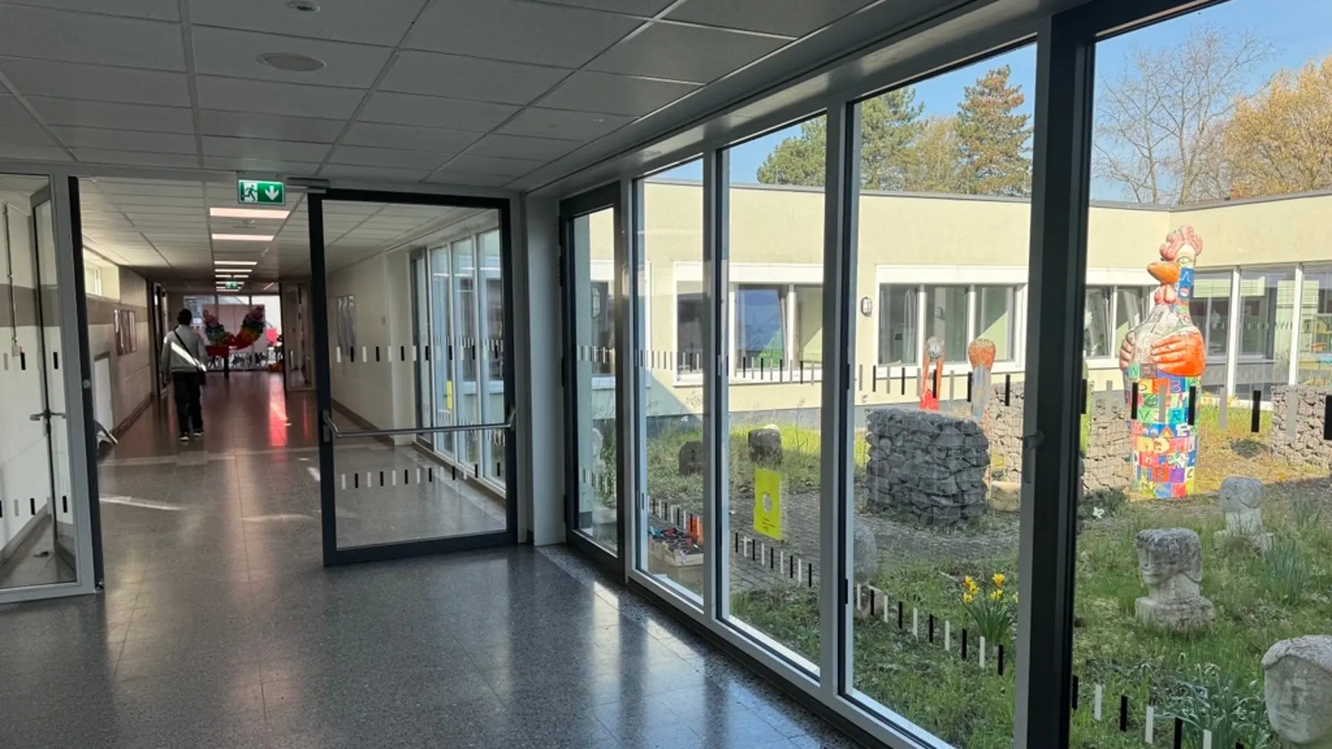 Ein heller, lichtdurchfluteter Flur in der Heinrich-Welsch-Schule. Eine bodentiefe Fensterfront gibt den Blick frei auf einen grünen Innenhof. Dort befinden sich verschiedene Kunstwerke, darunter Steinköpfe und eine große, bunte Hahnen-Skulptur.
