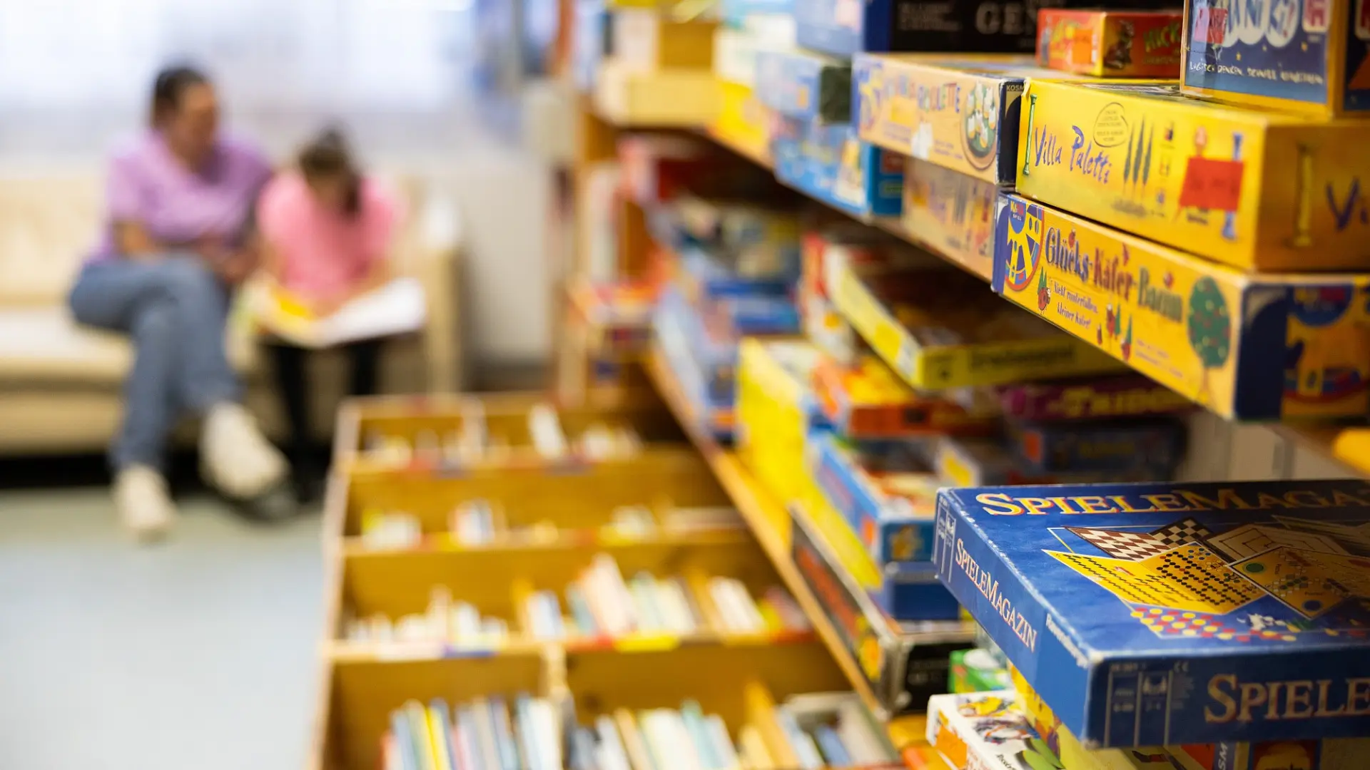 Wandregal voller Kinderbücher und -spiele, im Hintergrund eine erwachsene Person und ein Kind lesen am Sofa.