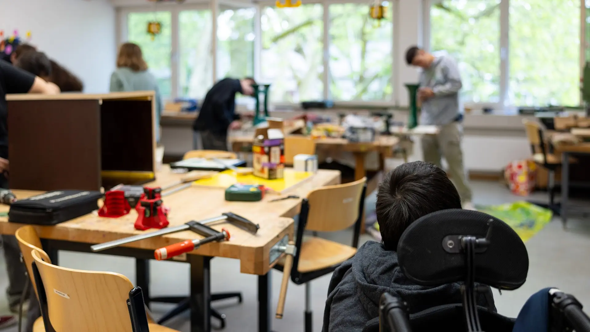 Eine Werkstatt mit Tischen mit Werkzeug, an denen Jugendliche arbeiten, vorne ist ein Kind im Rollstuhl.