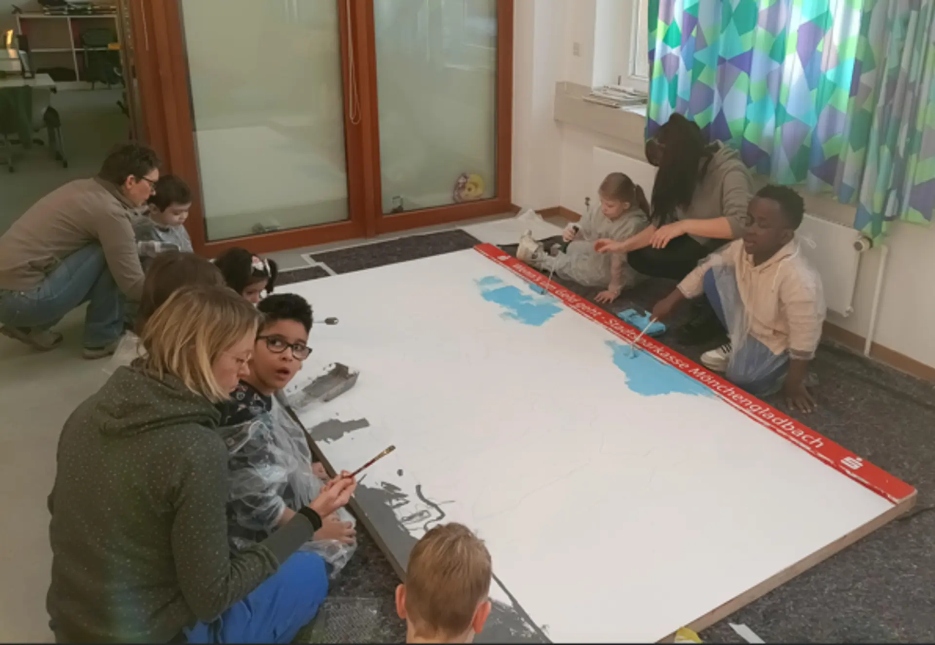 Mehrere Erwachsene und Kinder sitzen auf dem Boden um ein großes leeres Blatt und malen mit Pinseln darauf