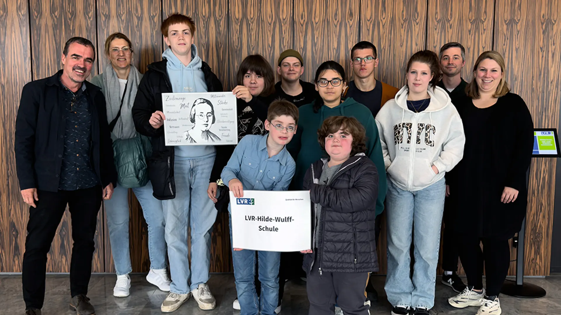Mehrere Personen (Erwachsene und Schüler*innen) stehen vor einer Wand und lächeln in die Kamera.