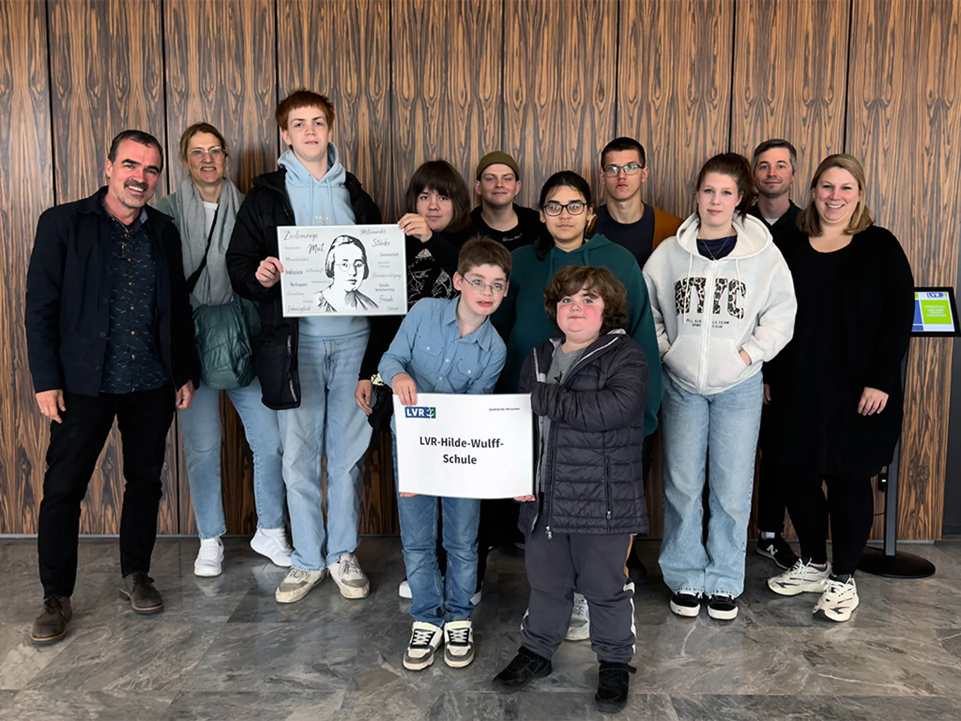 Mehrere Personen (Erwachsene und Schüler*innen) stehen vor einer Wand und lächeln in die Kamera.