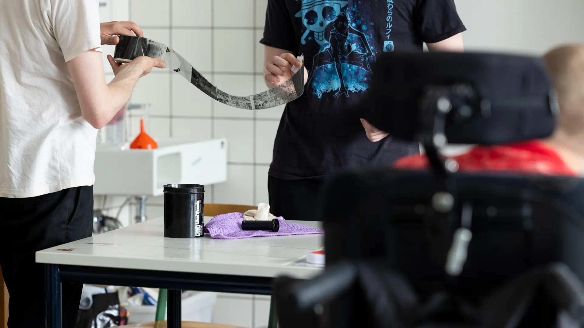 Zwei Personen halten eine entwickelte Analogfilmrolle, im Vordergrund ist ein Schulkind im Rollstuhl.
