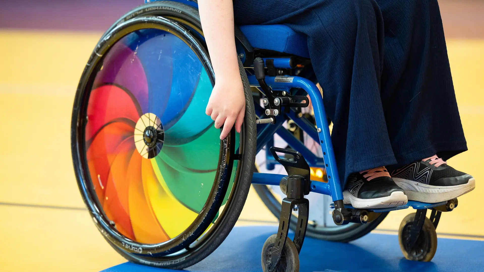 Ein Kind sitzt in einem Regenbogen-bunten Rollstuhl.