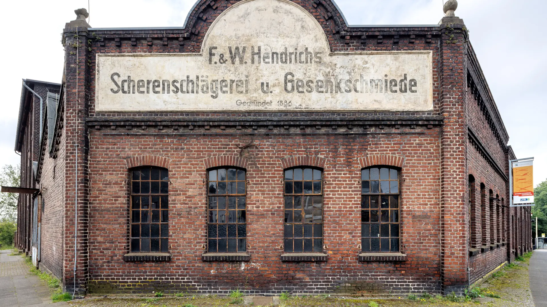 Ein roter Backsteinbau mit vier Fenstern. Über den Fenstern ist ein Schild installiert, auf dem steht: F & W Hendrichs Scherenschlägerei u. Gesenkschmiede.