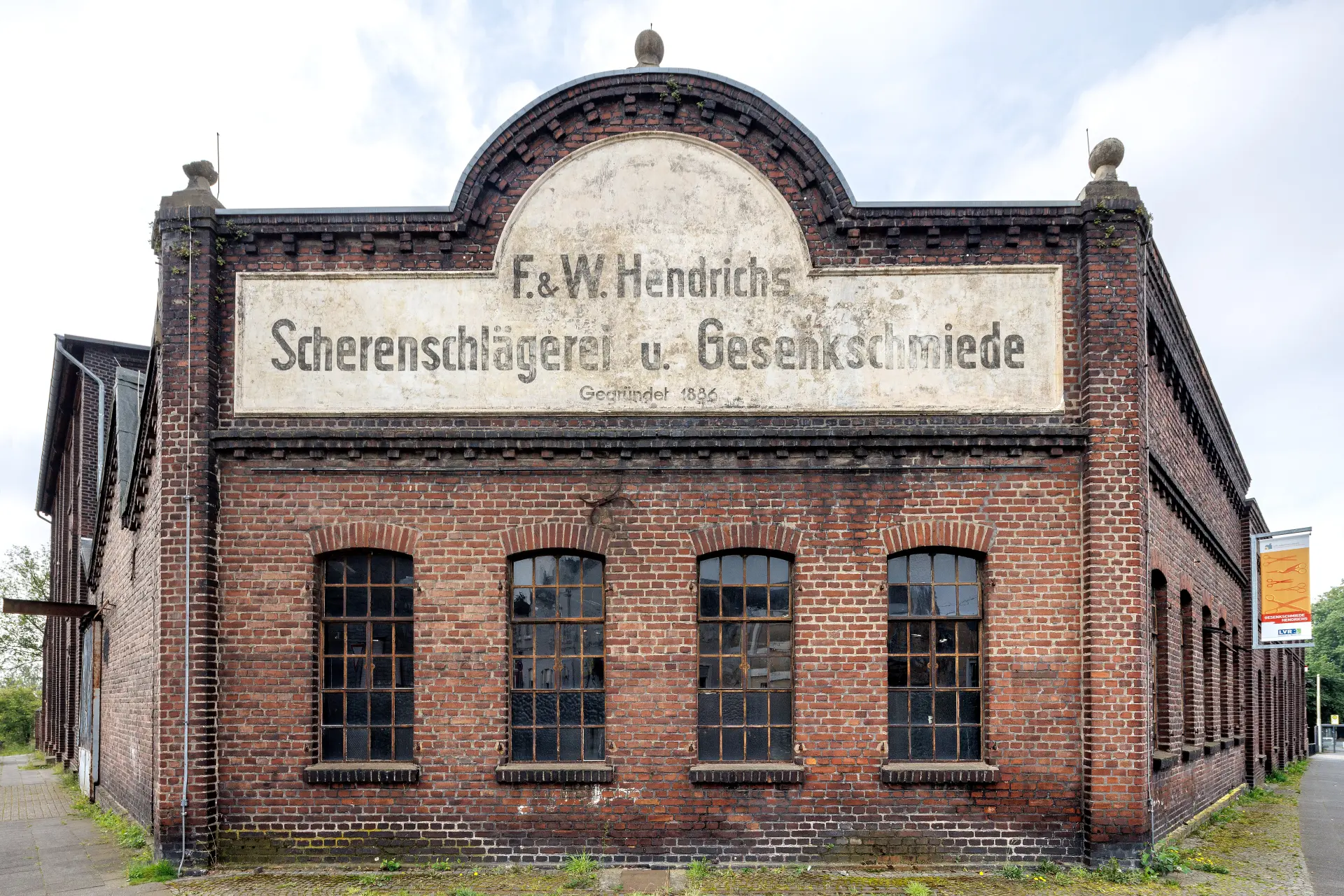 Ein roter Backsteinbau mit vier Fenstern. Über den Fenstern ist ein Schild installiert, auf dem steht: F & W Hendrichs Scherenschlägerei u. Gesenkschmiede.