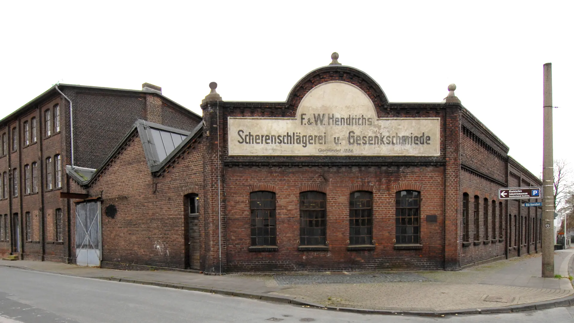 Ein Backsteingebäude an einer Straßenecke, auf dem ein Schild mit dem Namen "Gesenkschmiede Hendrichs" angebracht ist.