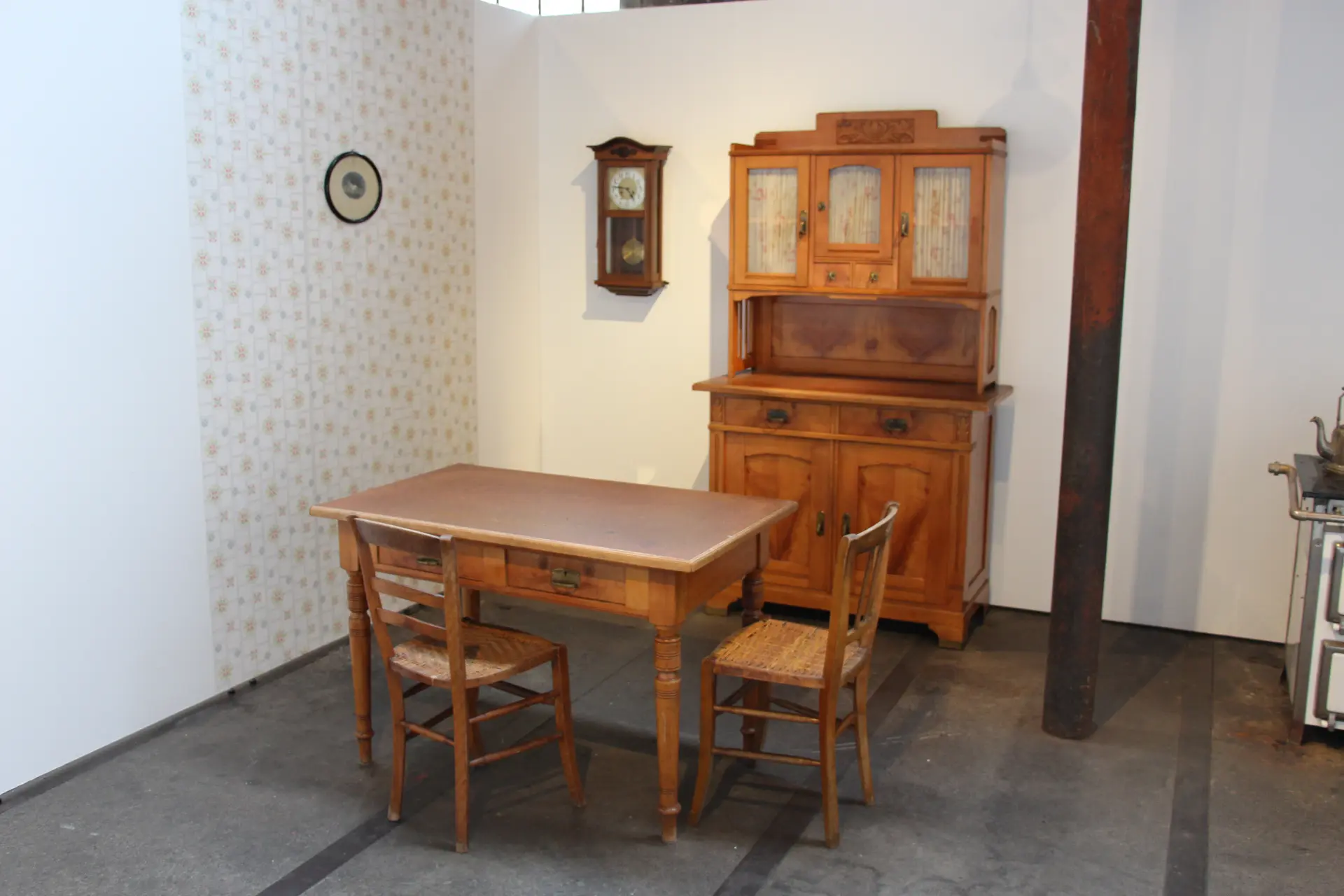 Ein Raum mit hellen Wänden und grauem Boden zeigt eine historische Wohnzimmereinrichtung. Im Mittelpunkt steht ein rechteckiger Holztisch mit zwei Stühlen aus Holz und geflochtener Sitzfläche. Dahinter befindet sich eine Anrichte mit Glaseinsätzen und dekorativen Elementen. An der Wand hängen eine Wanduhr und ein rundes Bild.