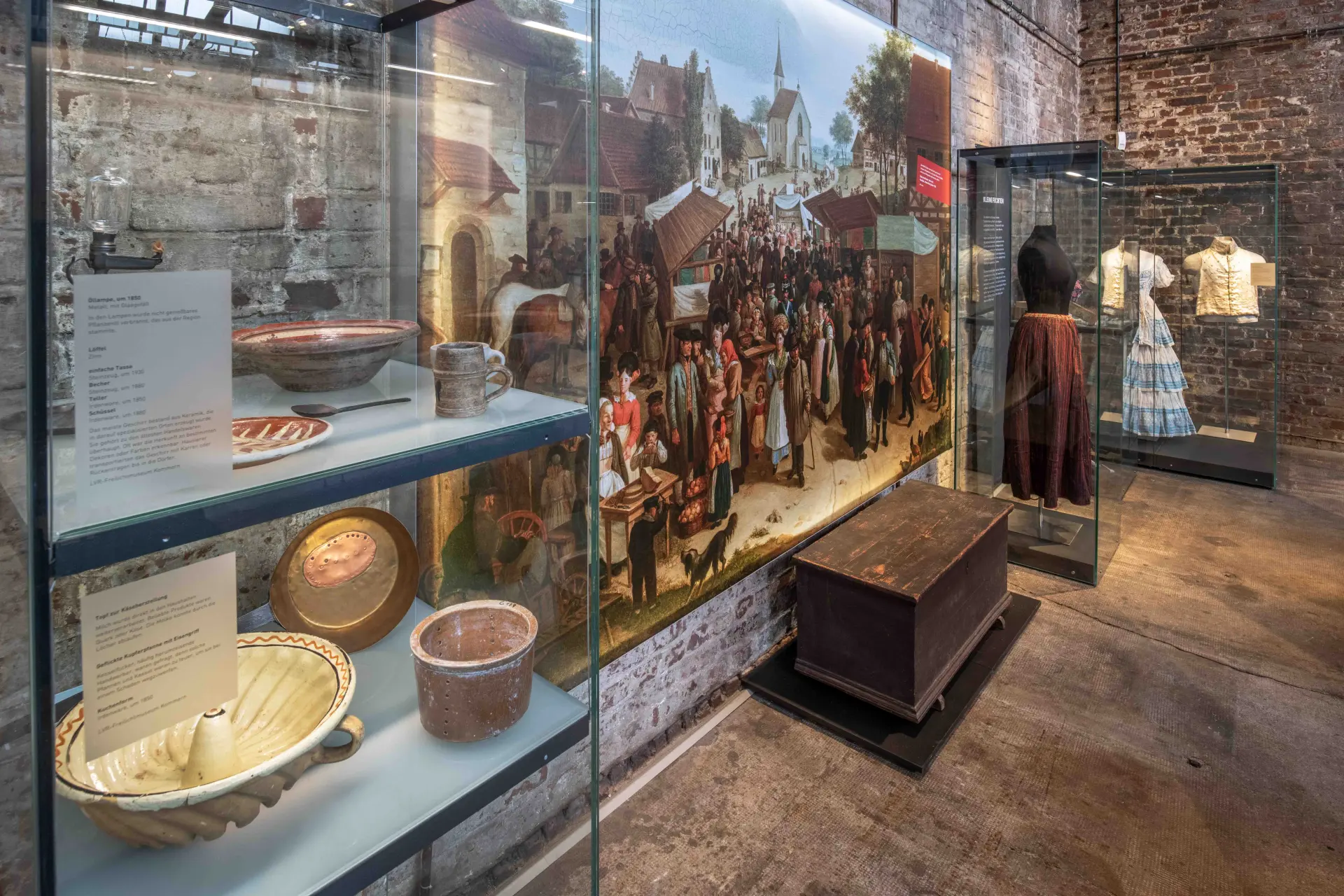 In einem Museumsraum stehen mehrere Glasvitrinen mit historischen Alltagsgegenständen wie Keramikschüsseln, Tellern und Besteck. An der Wand hängt ein großformatiges Gemälde, das eine belebte Marktszene mit vielen Menschen und Gebäuden zeigt. Daneben befindet sich eine große, dunkle Holztruhe. Auf der rechten Seite sind zwei historische Kleidungsstücke auf Figurinen ausgestellt, darunter ein Kleid mit blau-weißem Rock.
