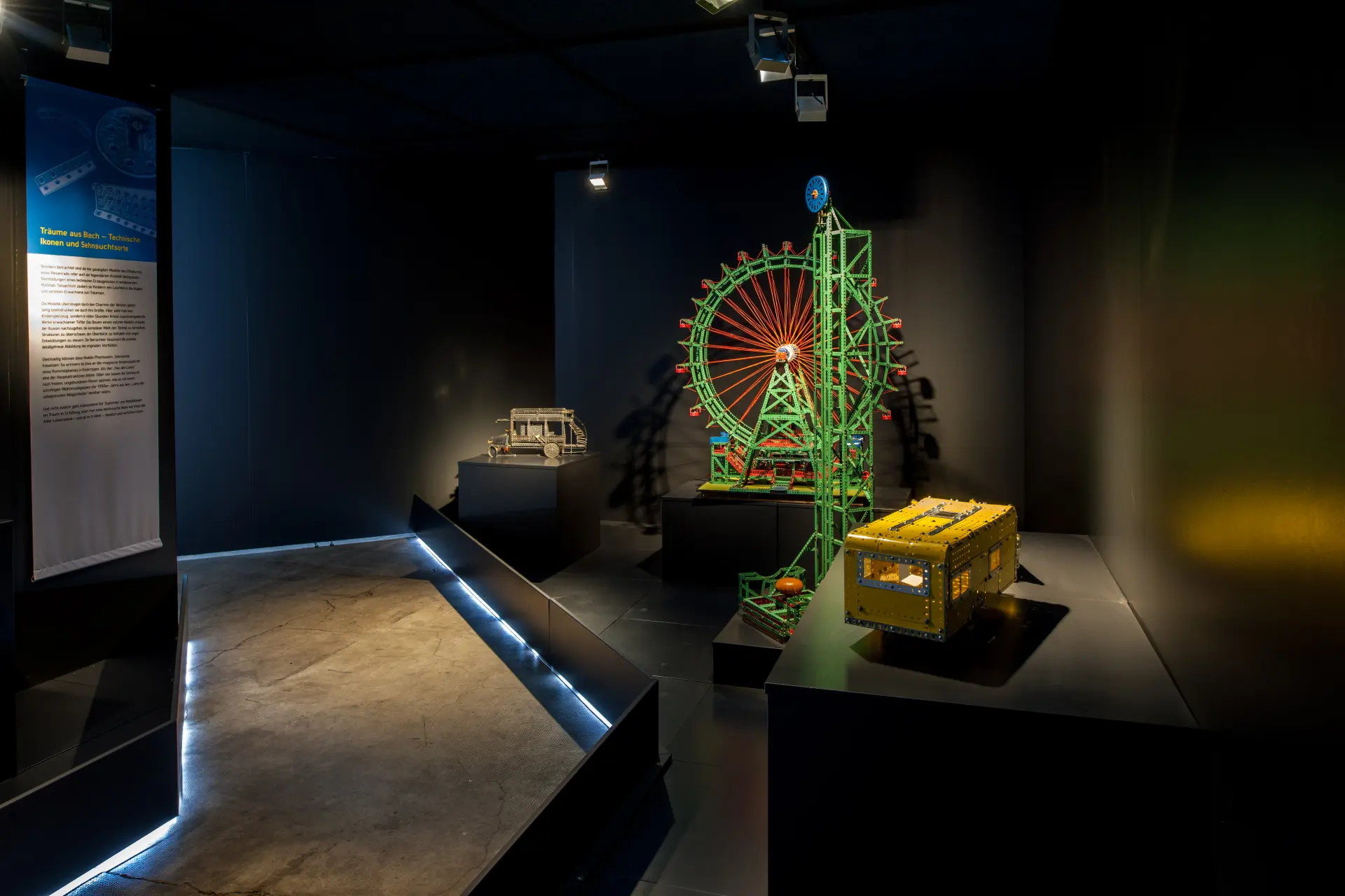 Dunkler Ausstellungsraum mit mehreren historischen Modellfahrzeugen und Maschinen. Im Mittelpunkt steht ein großes grünes Riesenradmodell mit roten Speichen, daneben ein gelbes Modellfahrzeug und ein kleines historisches Auto.