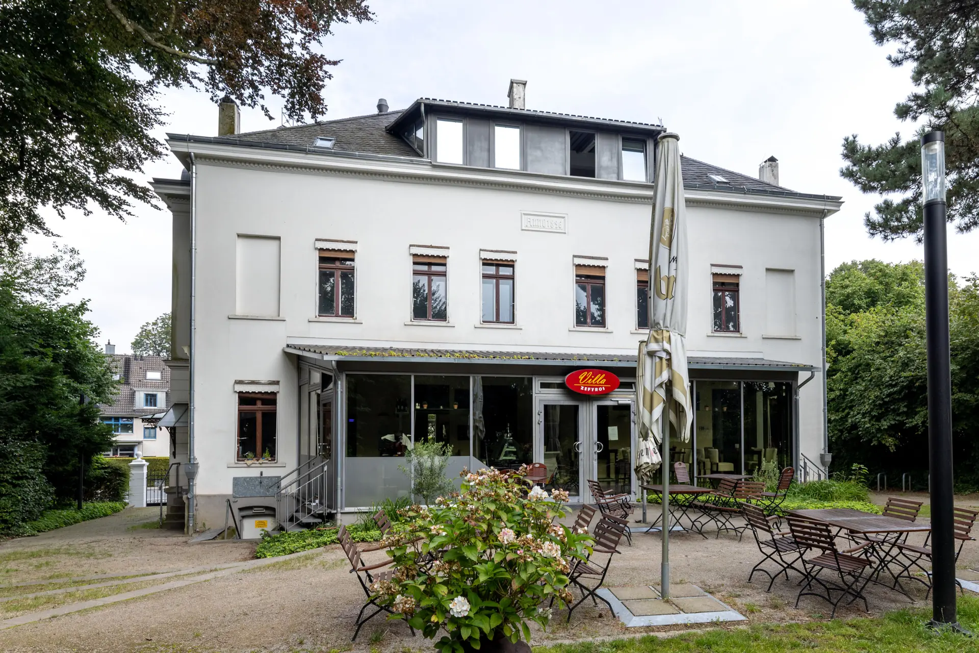 Eine weiße Villa mit Restaurant darin. Davor stehen Stühle und Tische in einem Außenbereich.