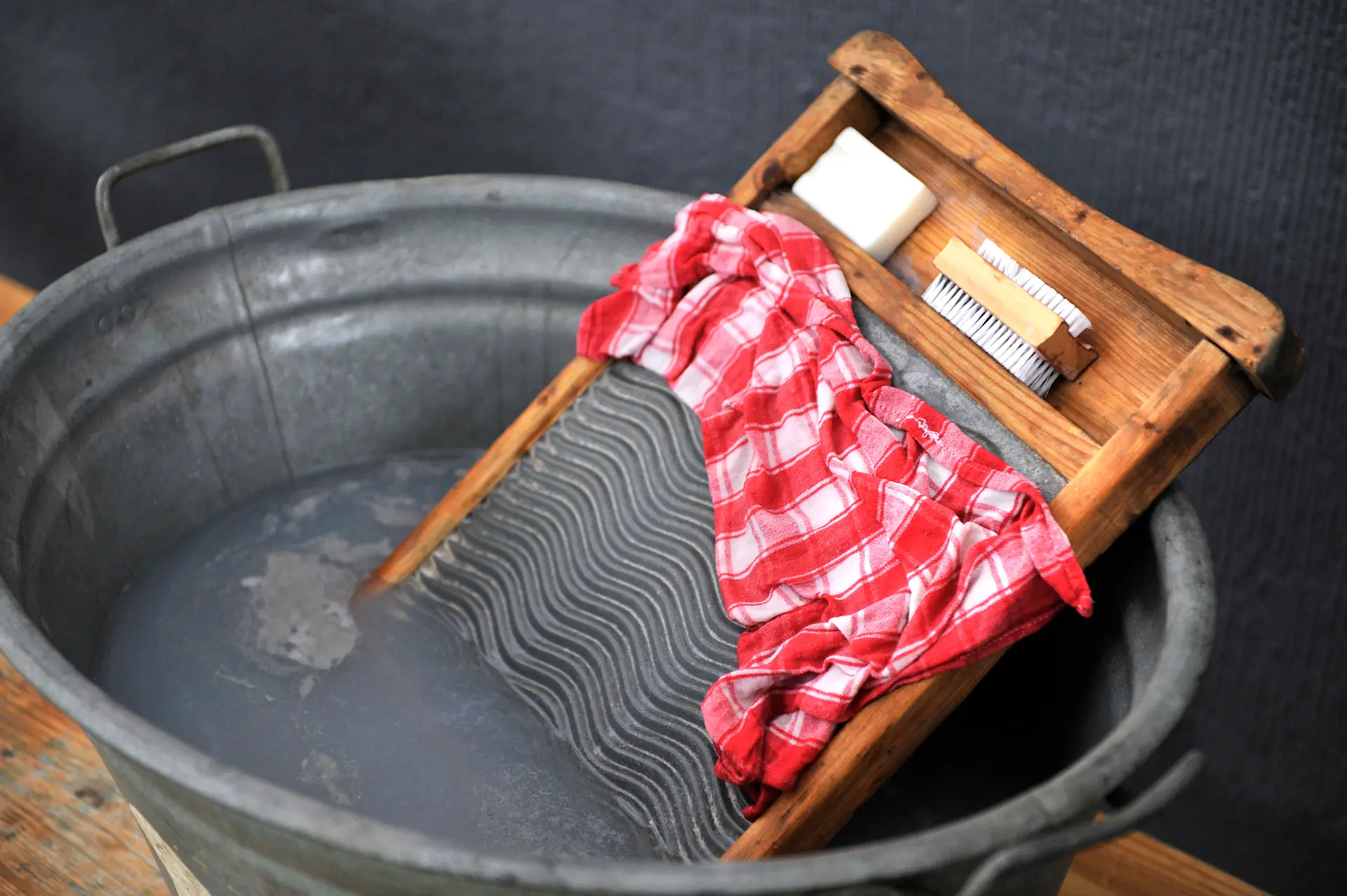 Ein Waschzuber gefüllt mit Wasser, in dem ein Waschbrett steckt. Auf dem Brett liegt ein nasses, rot-weiß kariertes Tuch, eine Bürste und ein Stück Seife.