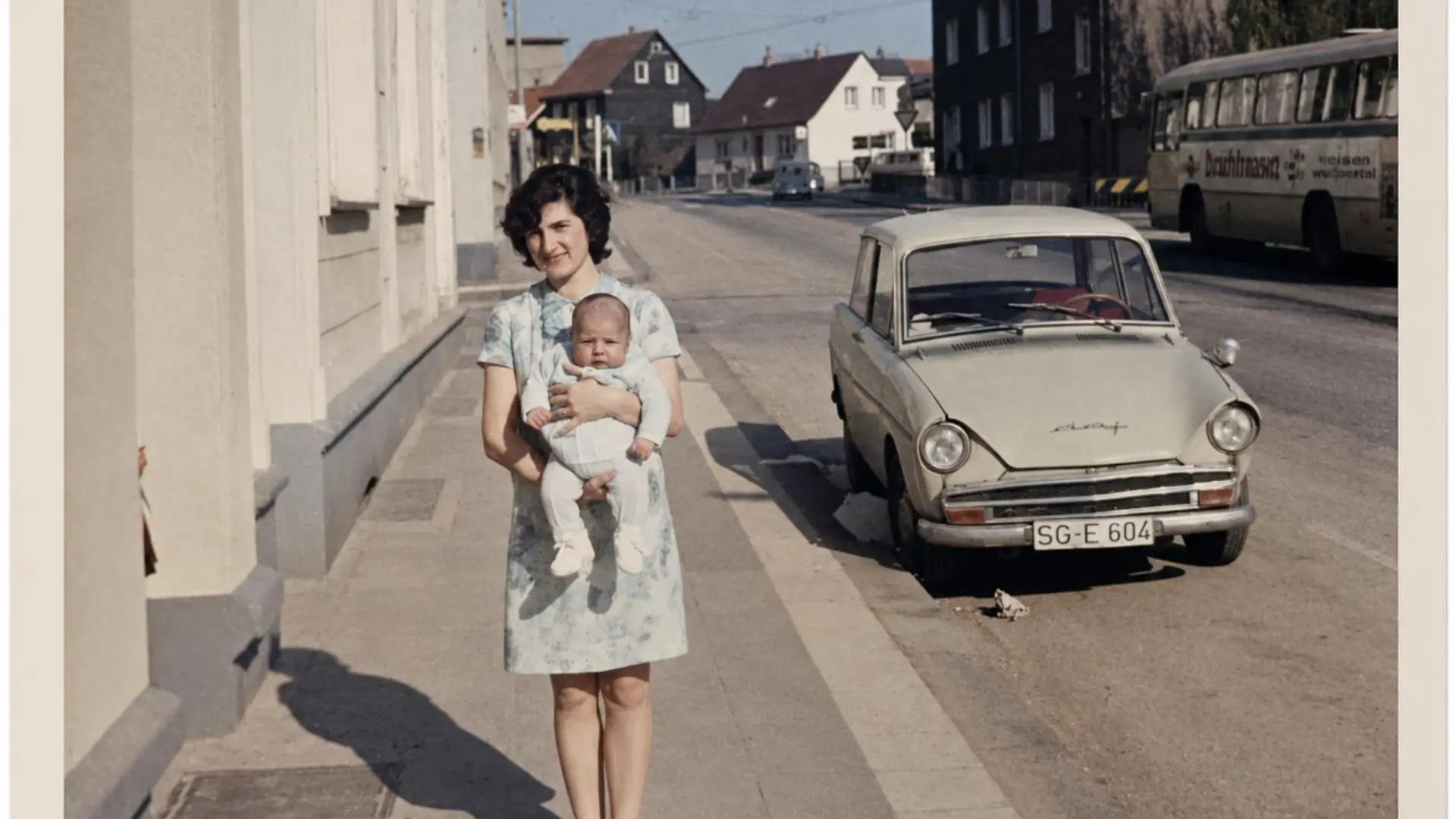 Frau in geblümtem Kleid hält Baby auf einem Bürgersteig neben einem geparkten Oldtimer-Kleinwagen mit deutschem Nummernschild