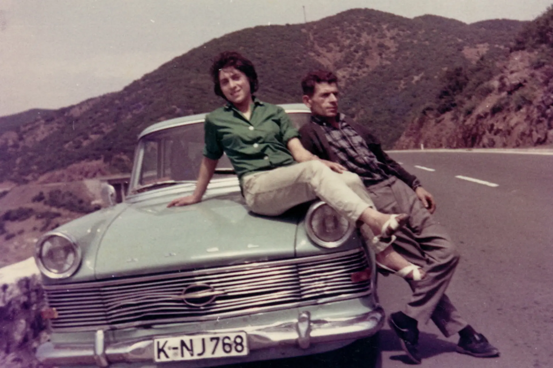 Zwei Personen lehnen an der Motorhaube eines alten Opel auf einer kurvigen Bergstraße.