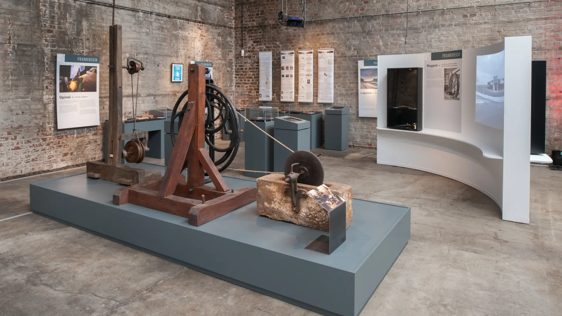Blick in einen Ausstellungsraum mit einer Schleifmaschine, Vitrinen auf Sockeln und einer Videowand.