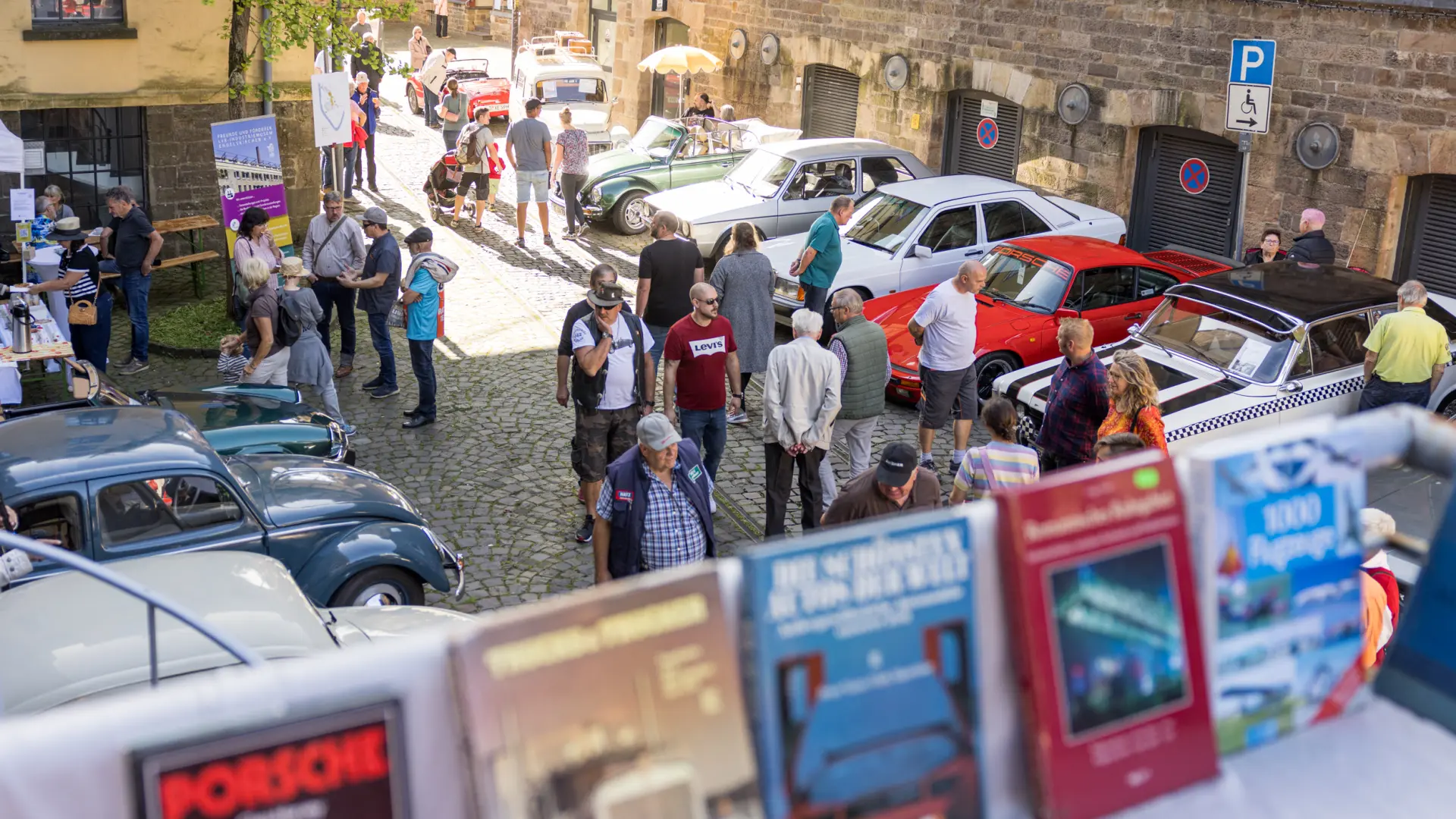 Blick über ausgestellte Automagazine auf einen Platz, auf dem Oldtimer stehen und Besuchende umhergehen.