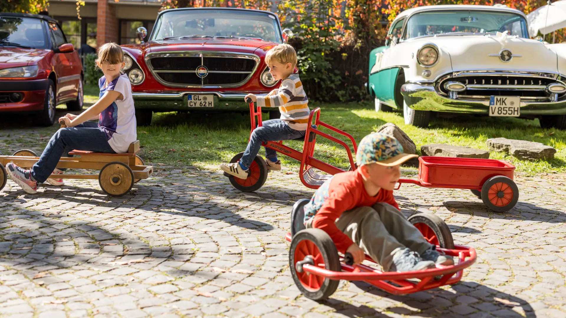 Einige Kinder mit Dreirädern vor historischen Sportwagen.