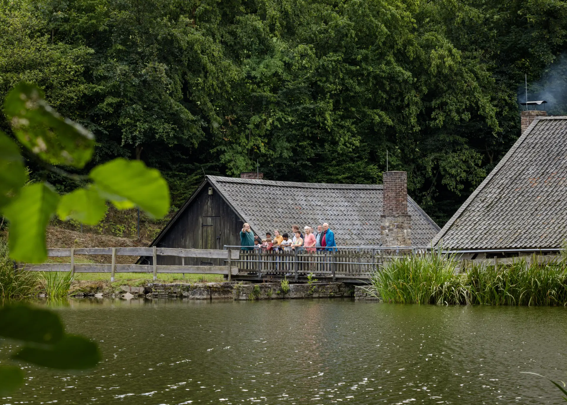 Außenansicht eines Gebäudes vor einem Teich, auf einem Steg steht eine Personengruppe