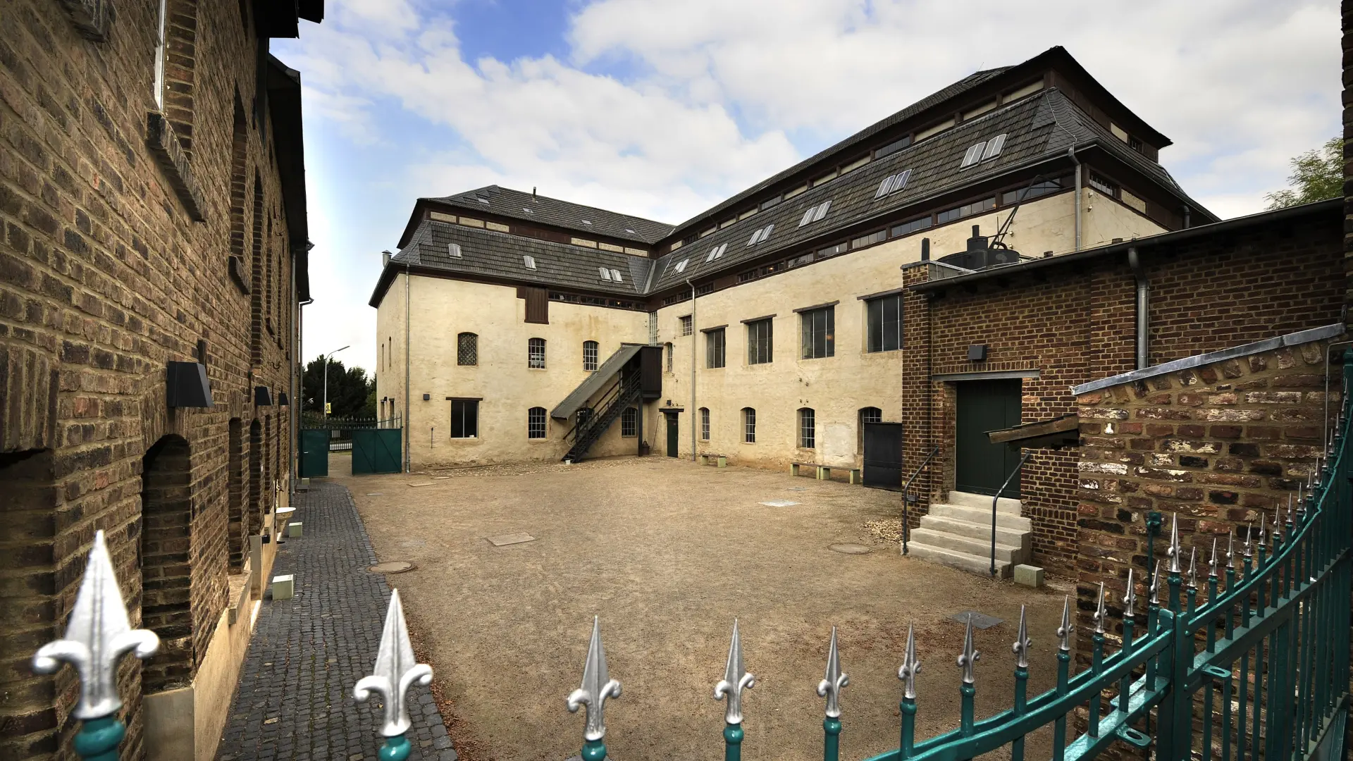 Blick über einen grünen Zaun auf einen Hof. Im Hintergrund ein gelbes Gebäude.