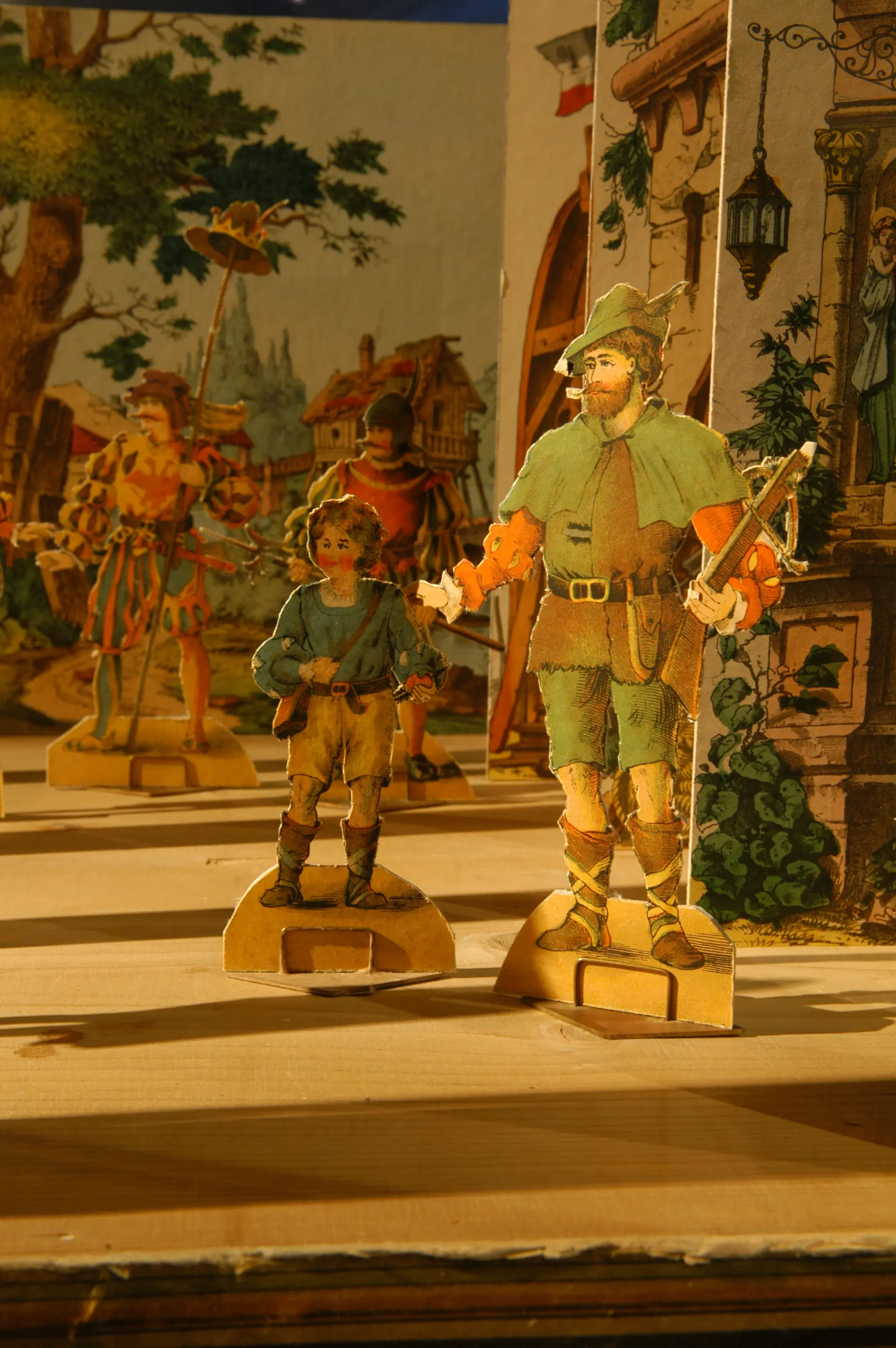 Historische Papierfiguren in mittelalterlicher Kleidung, die auf einer Holzfläche vor einer bemalten Kulisse mit Wald, Häuser und einer Burg stehen.