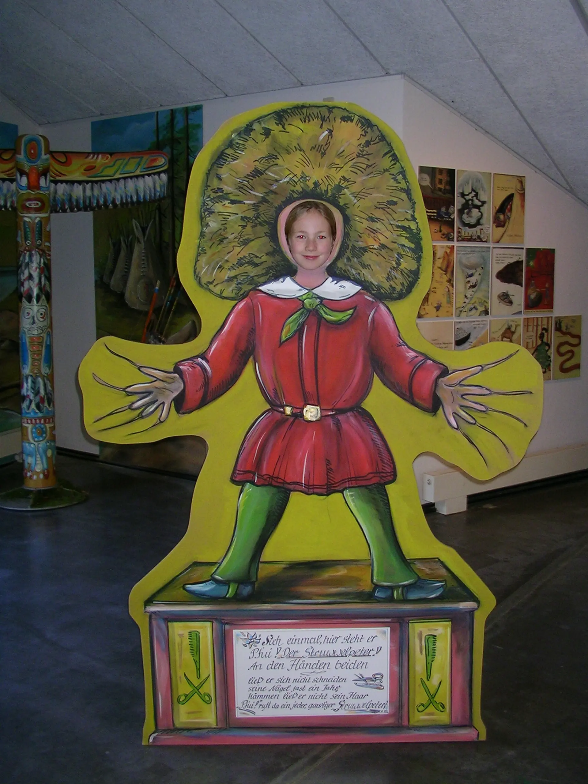 Ein Kind steht hinter einer Figur mit roter Kleidung, grünen Strümpfen, langen Fingernägeln und wuscheligen Haaren. das Kind steckt seinen Kopf durch die Figur hindurch.