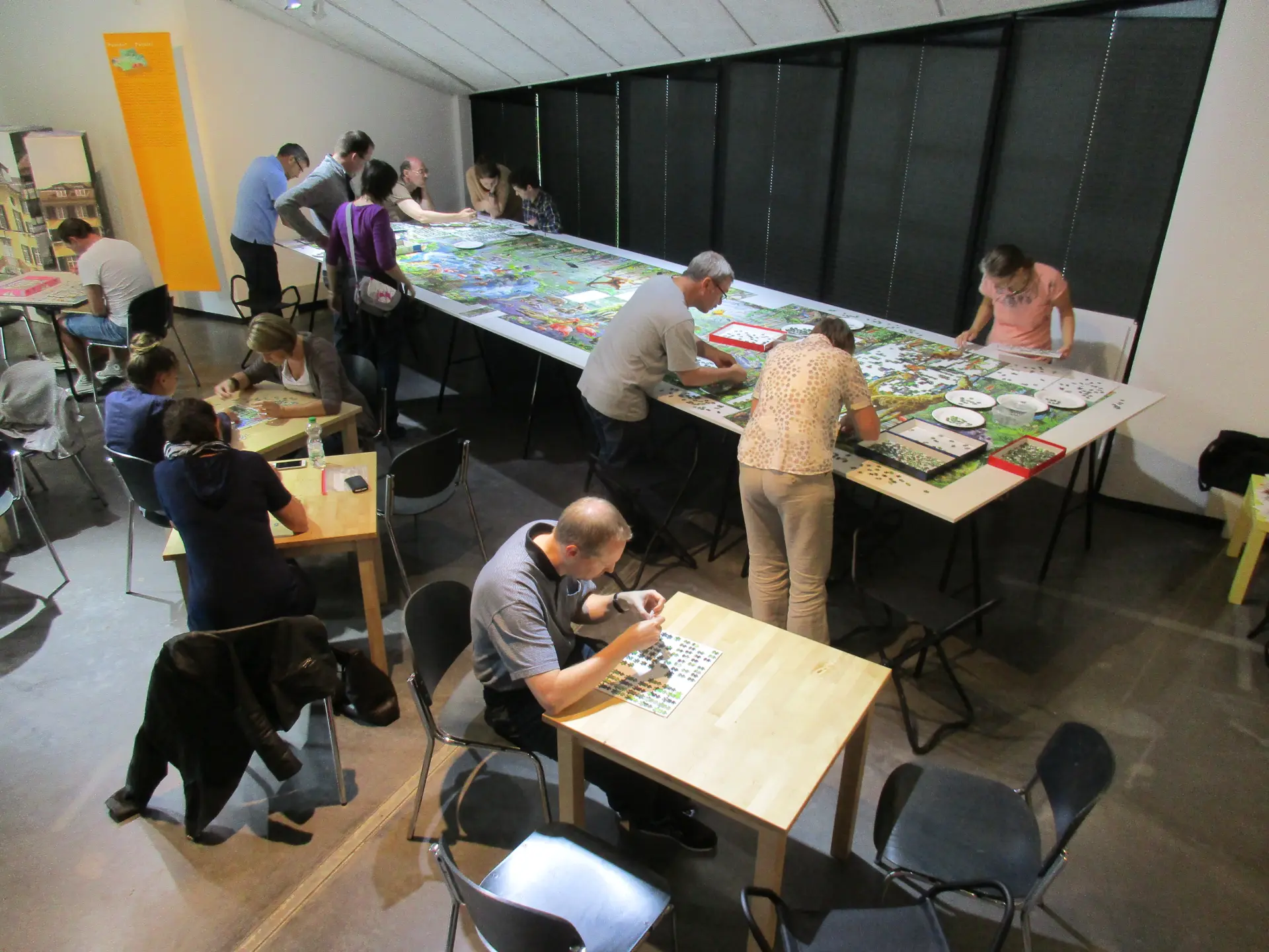 Blick in einen Raum, in dem Menschen an mehreren Tischen sitzen und Puzzle machen. Im Hintergrund stehen einige Menschen um einen großen Tisch und puzzeln an einem großen Puzzle.